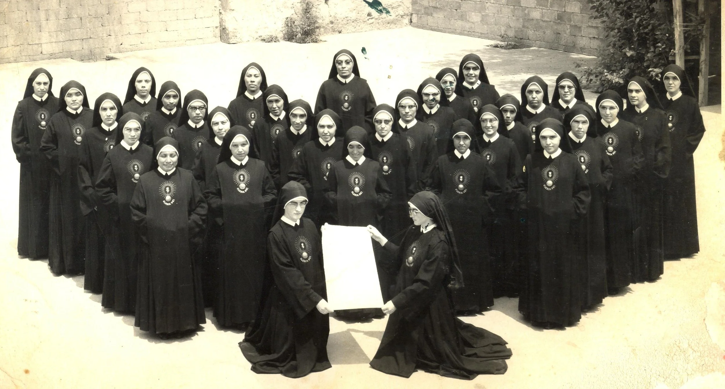 Grupo de religiosas vestidas con hábito negro y velo, posando para una fotografía, con dos religiosas en el primer plano sosteniendo un marco o cartel.