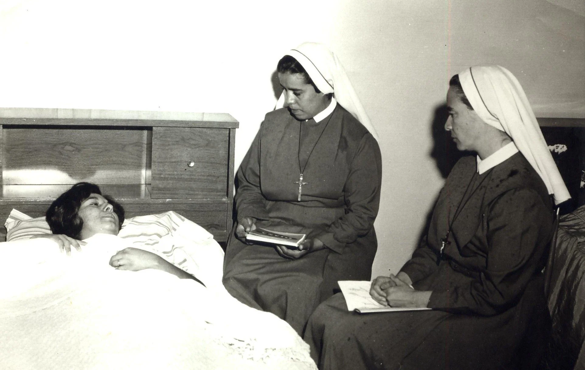 Dos monjas están hablando con una mujer enferma en una cama en un entorno hospitalario, una de las monjas está tomando notas.
