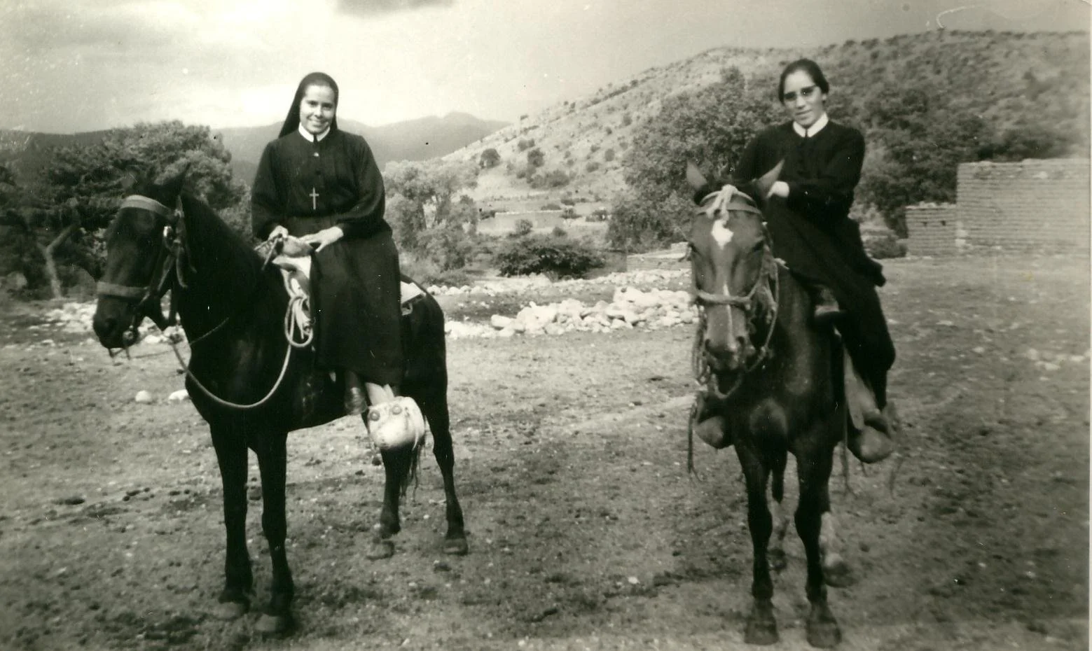 Dos monjas montando caballos en un paisaje rural con colinas y árboles.