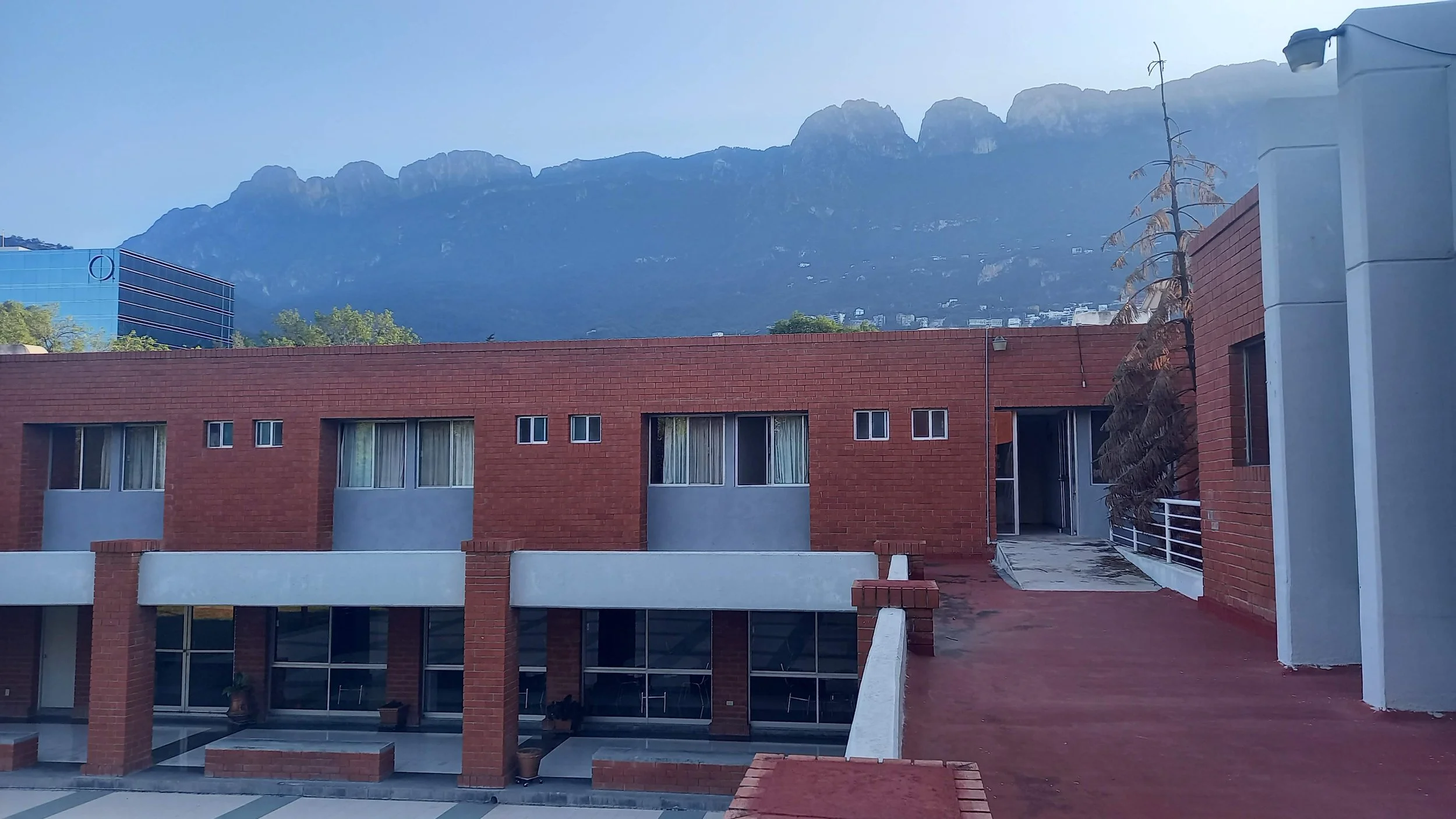 edificio moderno de ladrillo con ventanas y un patio en la parte inferior, fondo montañas y cielo despejado.