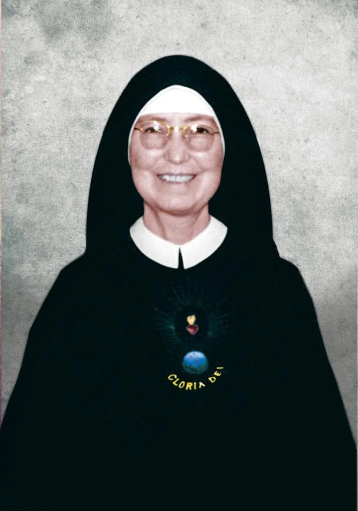 Imagen de una monja con expresión sonriente, usando hábito tradicional con un medallón en el pecho y fondo gris.