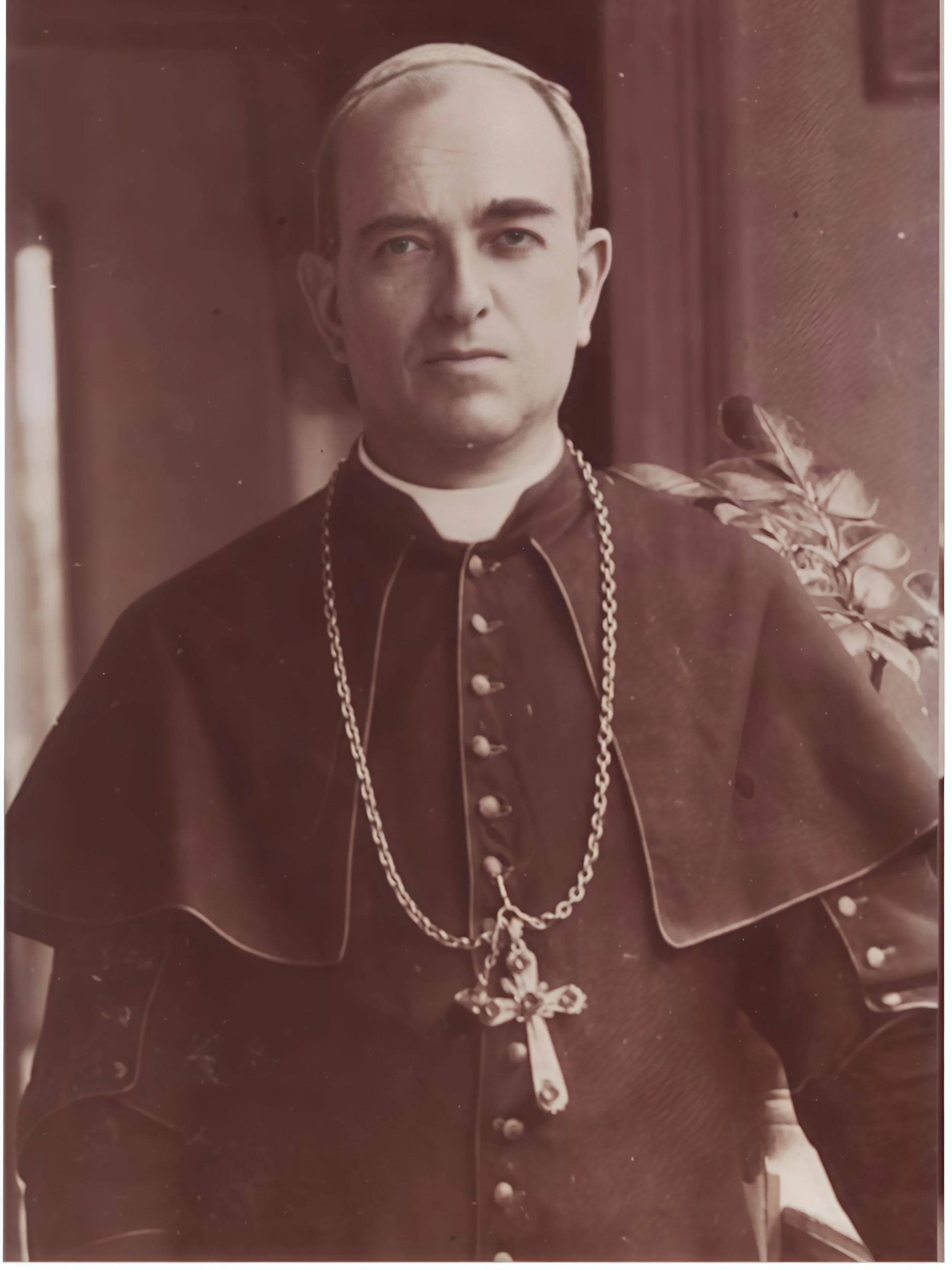 Foto en sepia de un hombre vestido con ropa clerical, llevando una cadena con una cruz grande y un rostrillo, posando frente a un fondo con plantas