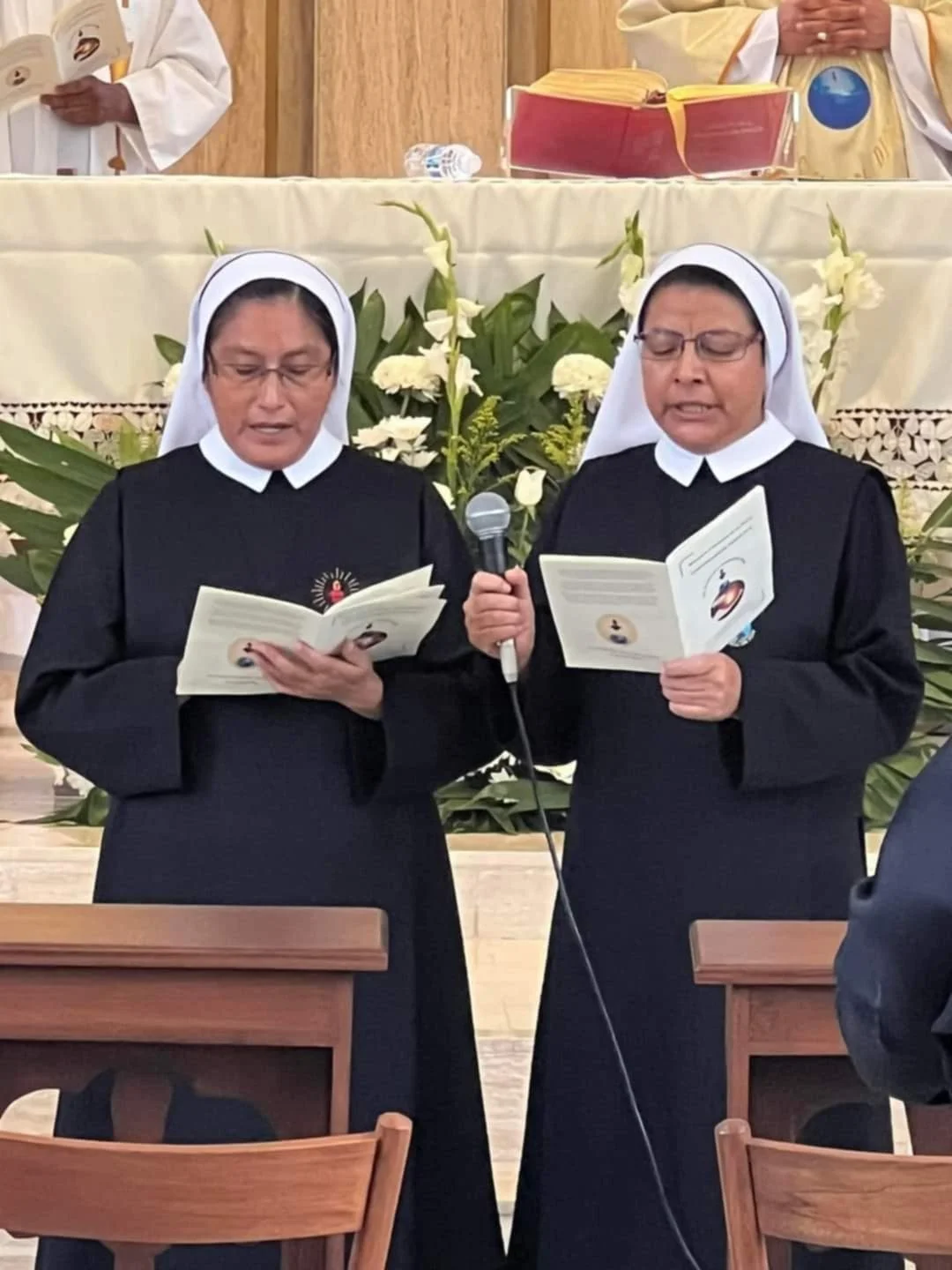 Dos monjas vestidas de hábito negro y velo blanco rezando y leyendo libros en un entorno religioso.