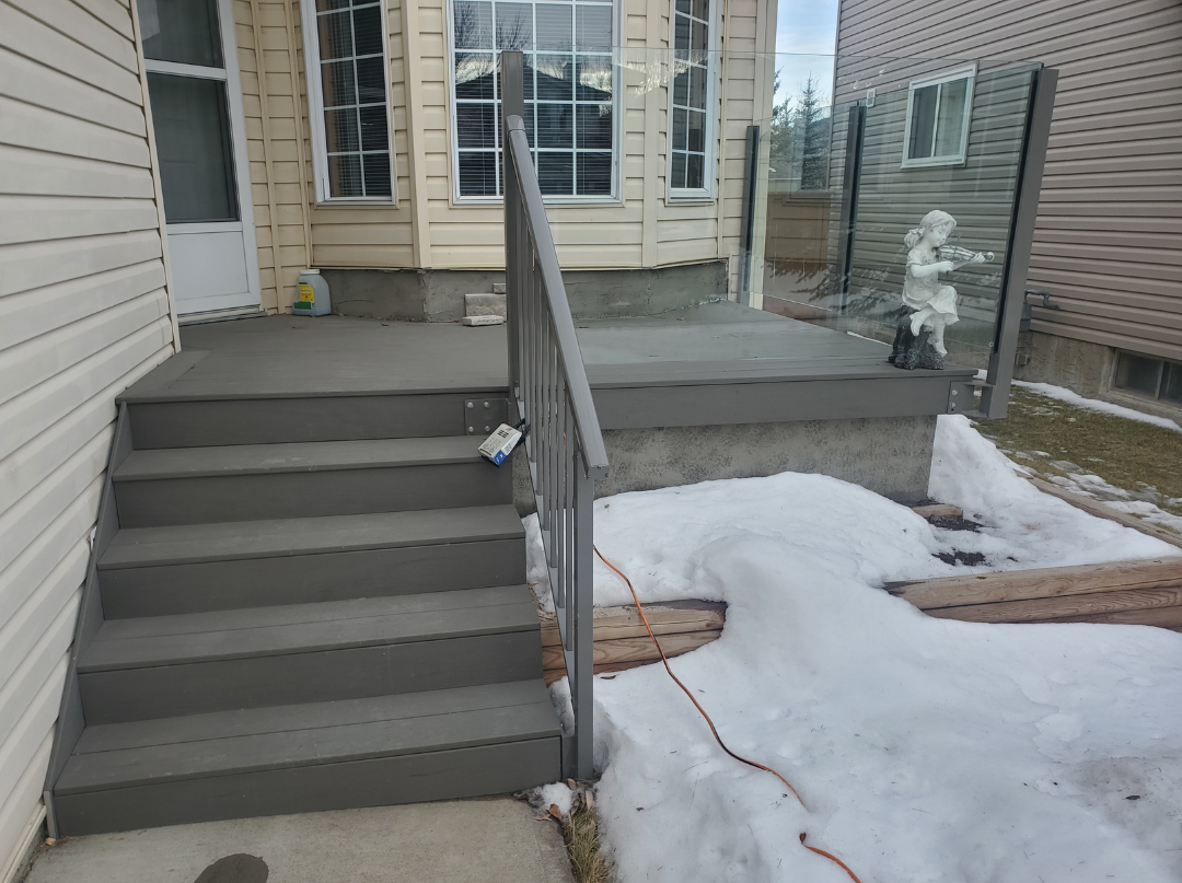 Gray wooden stairs leading to a back door with a small porch, surrounded by snow and a garden hose, with a glass railing featuring a decorative statue of a girl sitting and playing a flute.