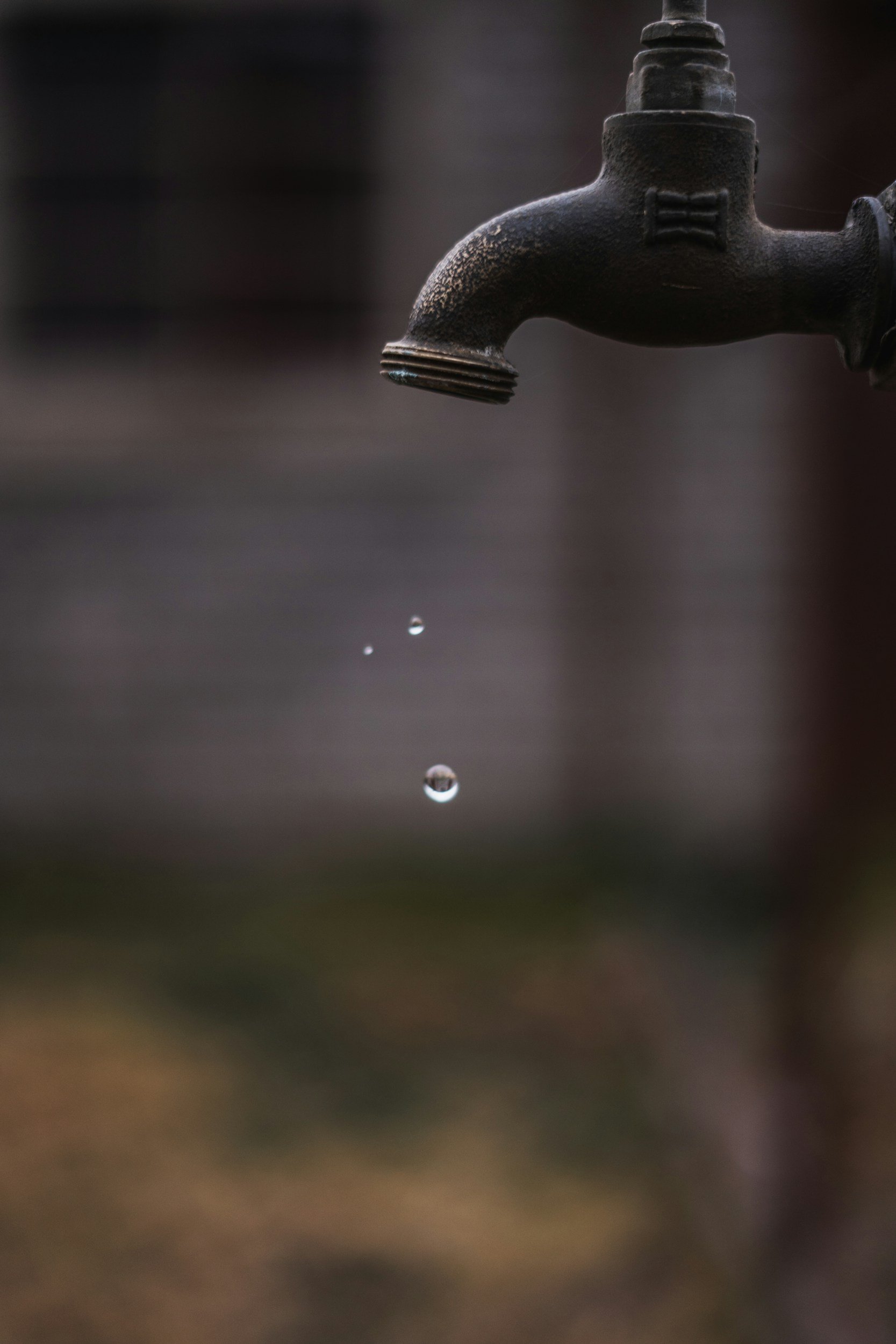 Close-up of an old, black outdoor faucet with water droplets falling from its spout against a blurred background.