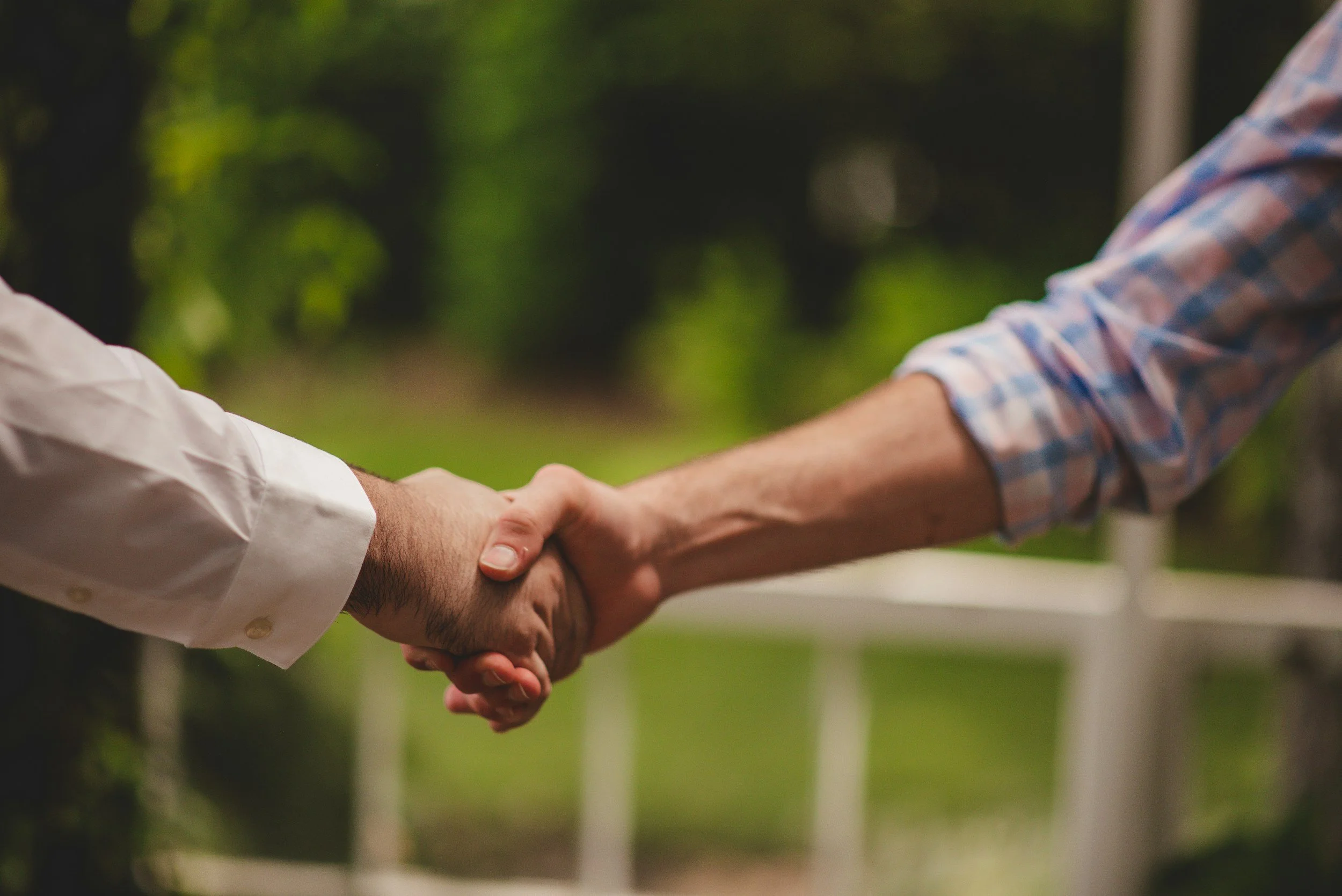 Two people shaking hands outdoors, one wearing a white shirt and the other wearing a checkered shirt.