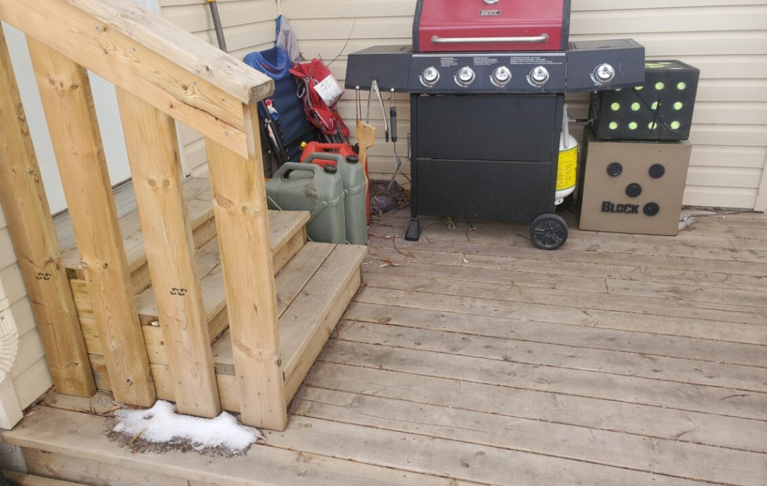 Outdoor wooden deck with a black barbecue grill, a set of concrete blocks with green circular holes, a propane tank, watering cans, bags, and gardening tools. The deck has some snow at the edge.