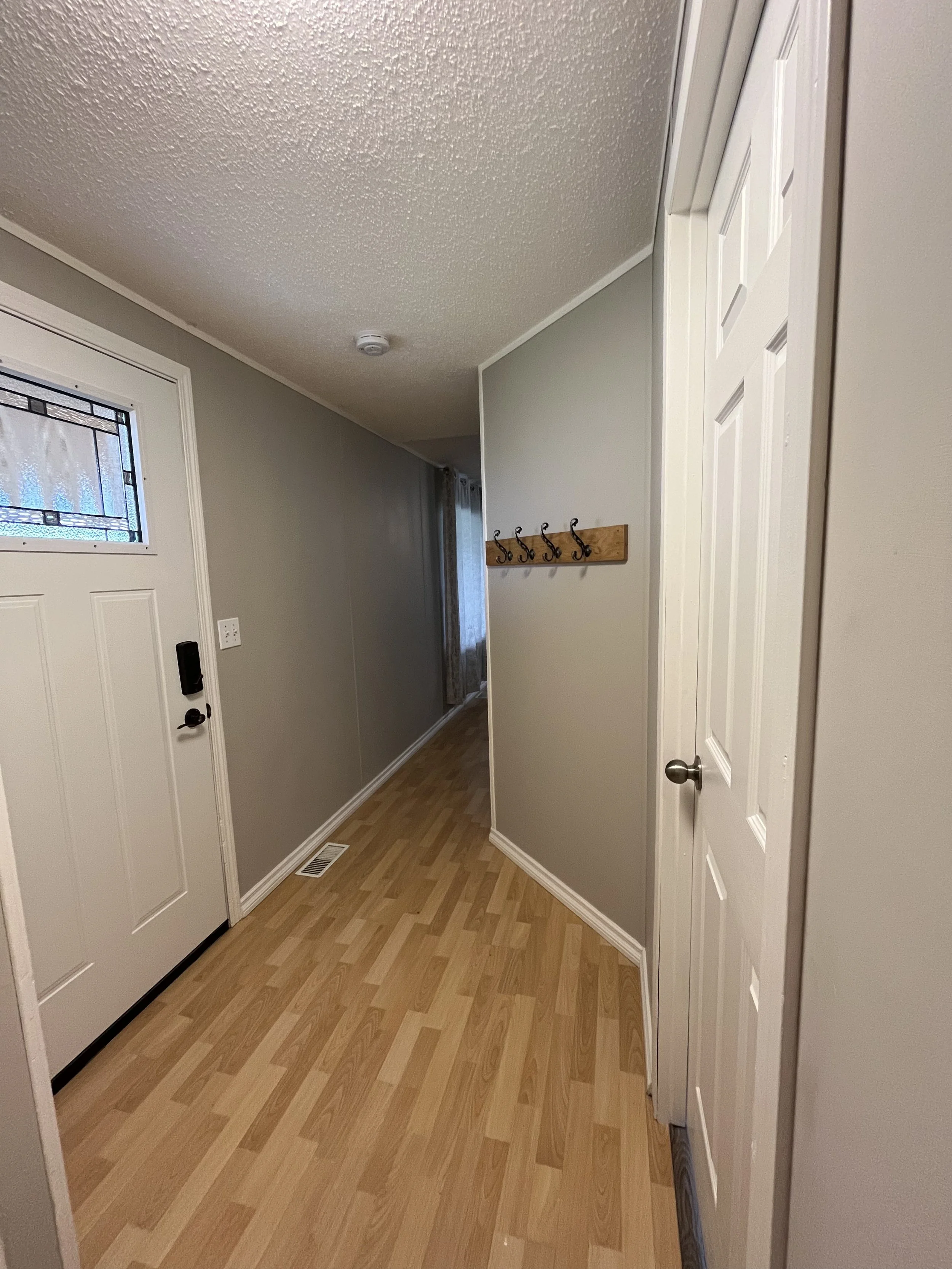 Entryway with front door, small window, wooden coat rack, light gray walls, wood laminate flooring, and a doorway leading to another room.
