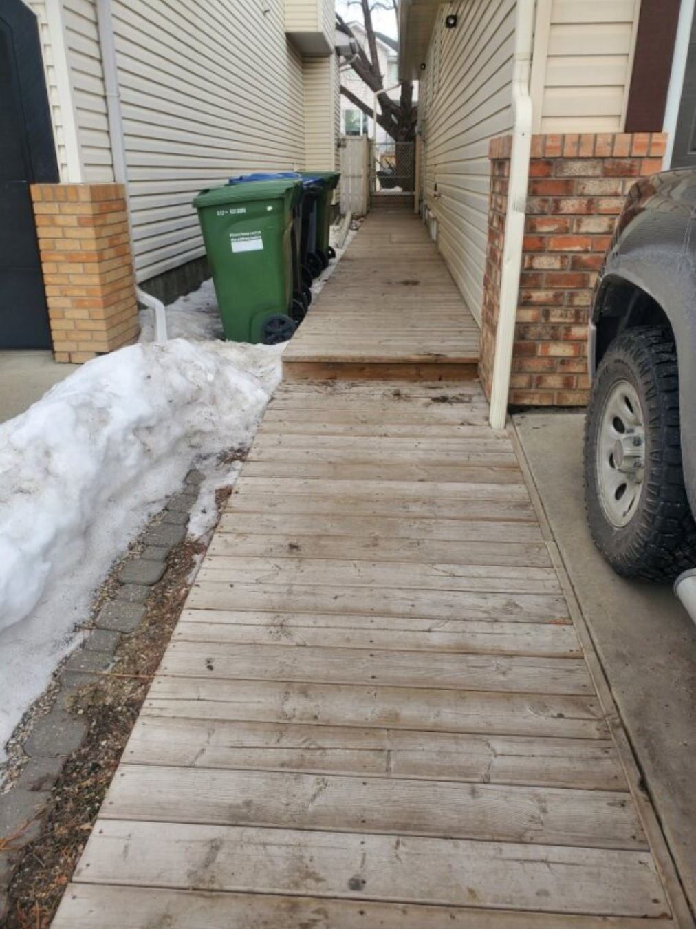 Side alley with a wooden walkway, snow on the ground, green and blue trash bins, side of a house with siding and brick, part of a car parked nearby, and a tree visible in the background.