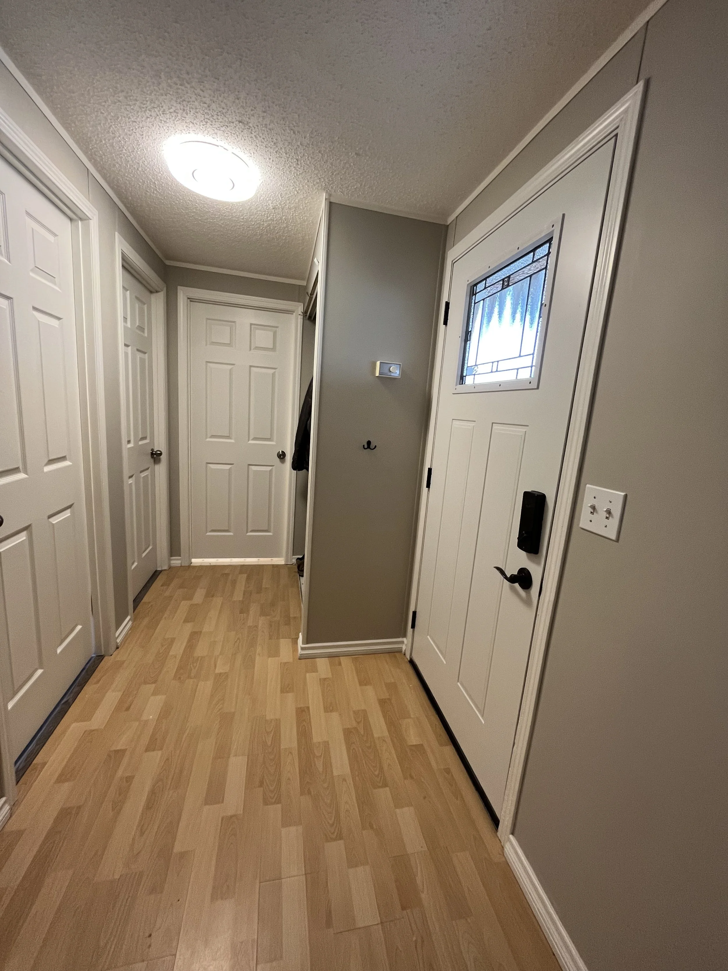 An entryway with a white front door with a small window, a lock, and a handle. Light-colored hardwood flooring, gray walls, with multiple closet doors on the left side, a thermostat on the wall, and a ceiling light fixture.