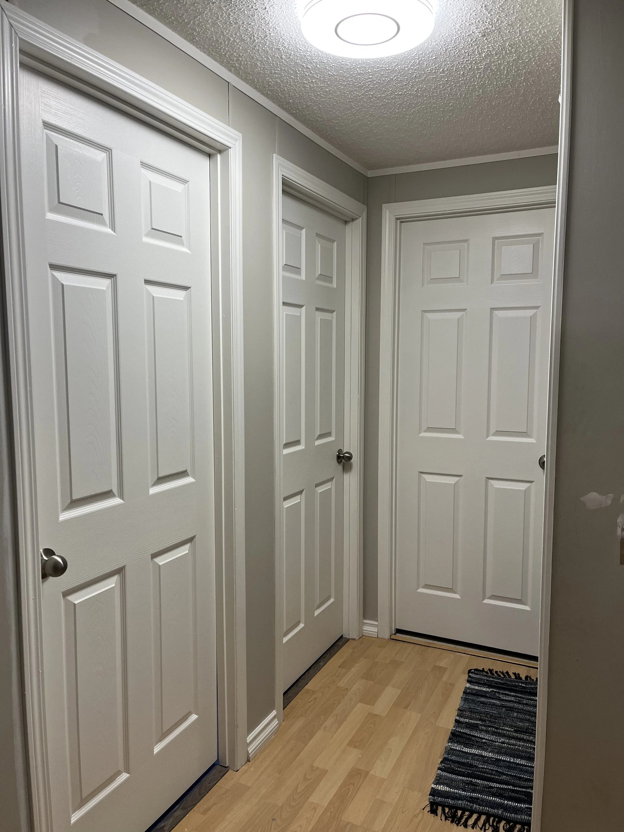 A hallway with three white interior doors, a ceiling light fixture, light-colored wooden flooring, and a small black and white striped rug.