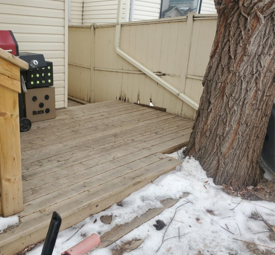 A wooden deck next to a house with snow on the ground, a tree trunk growing through the deck, and a grill on the left side.