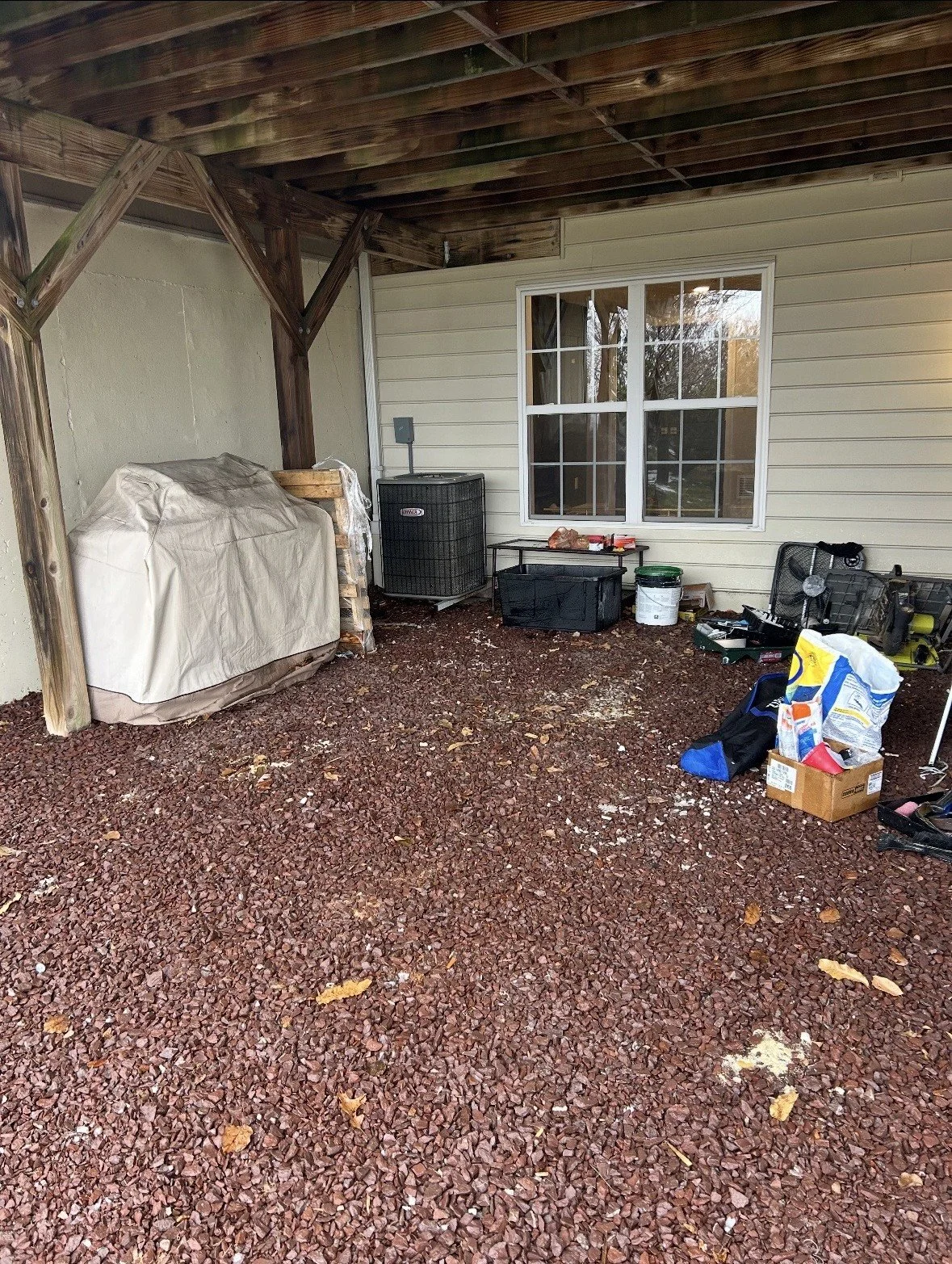 Partially covered backyard patio with storage boxes, tools, and miscellaneous items on a gravel ground, adjacent to a house with a large window.