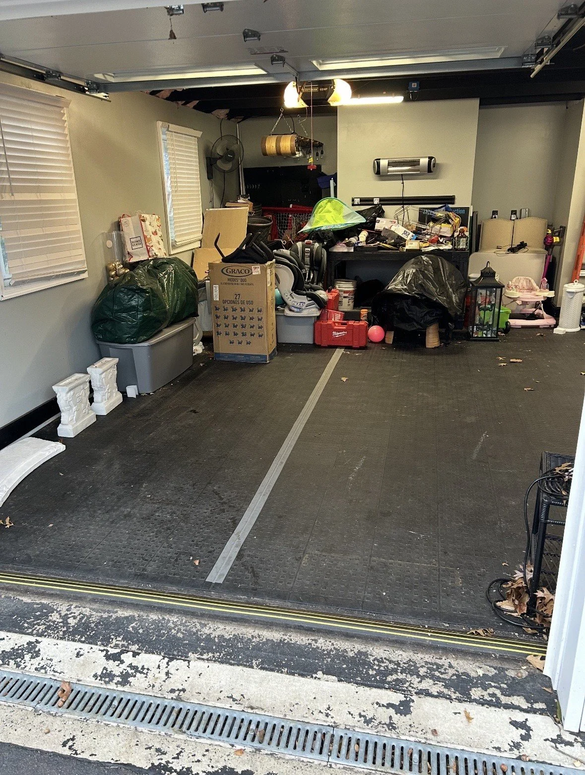 A cluttered garage with items like boxes, bags, and tools scattered around, and the garage door open to the outside driveway.