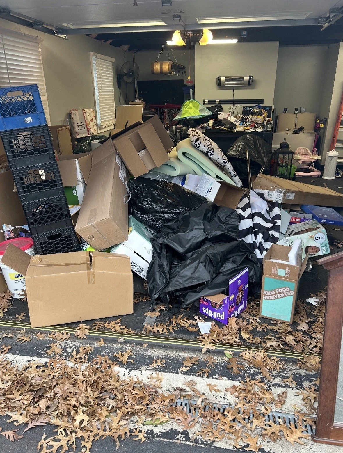 A cluttered garage or storage space with cardboard boxes, plastic bags, foam mats, and various household items scattered on the floor and covered surfaces.