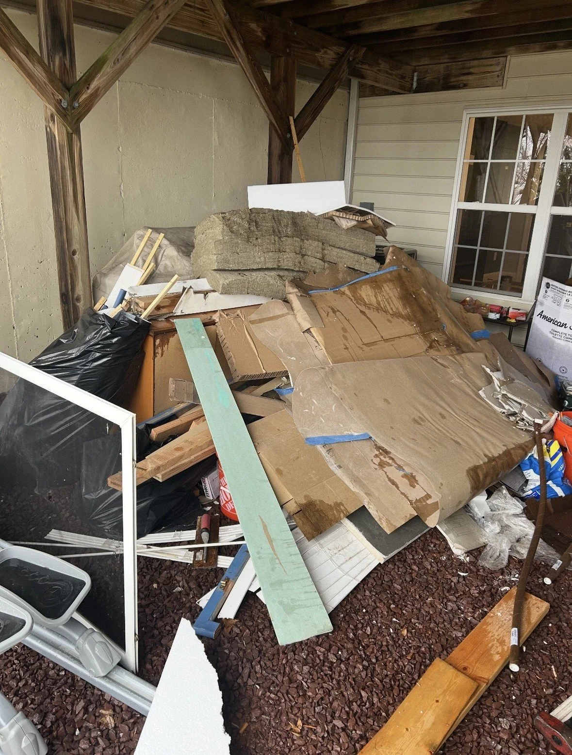 A cluttered outdoor porch area with construction debris, cardboard, and insulation materials stacked against the wall under a wooden roof.