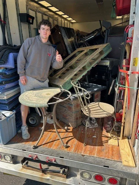 A young man stands inside a moving truck filled with furniture and household items, giving a thumbs-up gesture.
