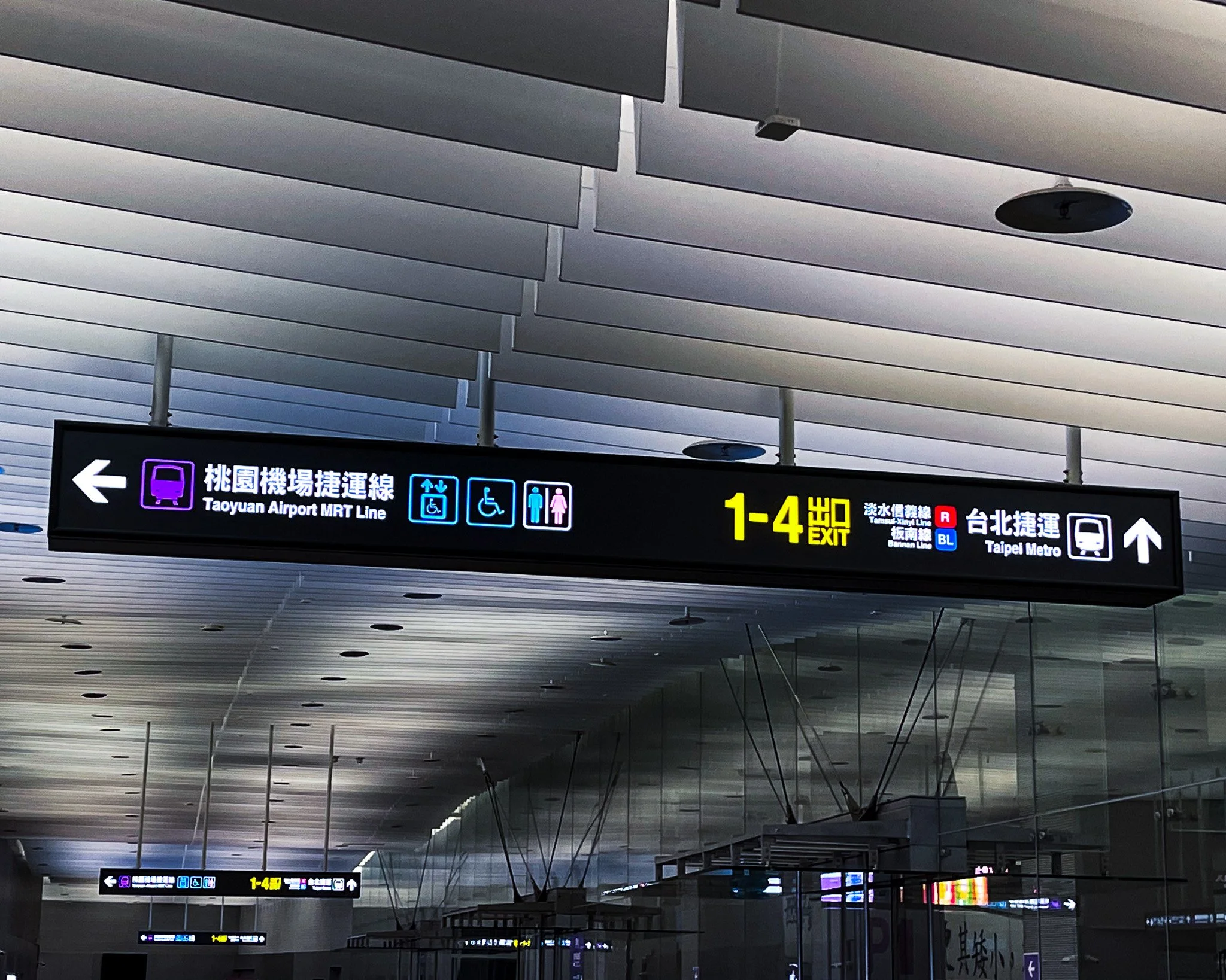 Signboard at an airport showing directions to the Taoyuan Airport MRT Line and Taipei Metro, with icons for accessible facilities and restrooms, and indicating exits 1-4.