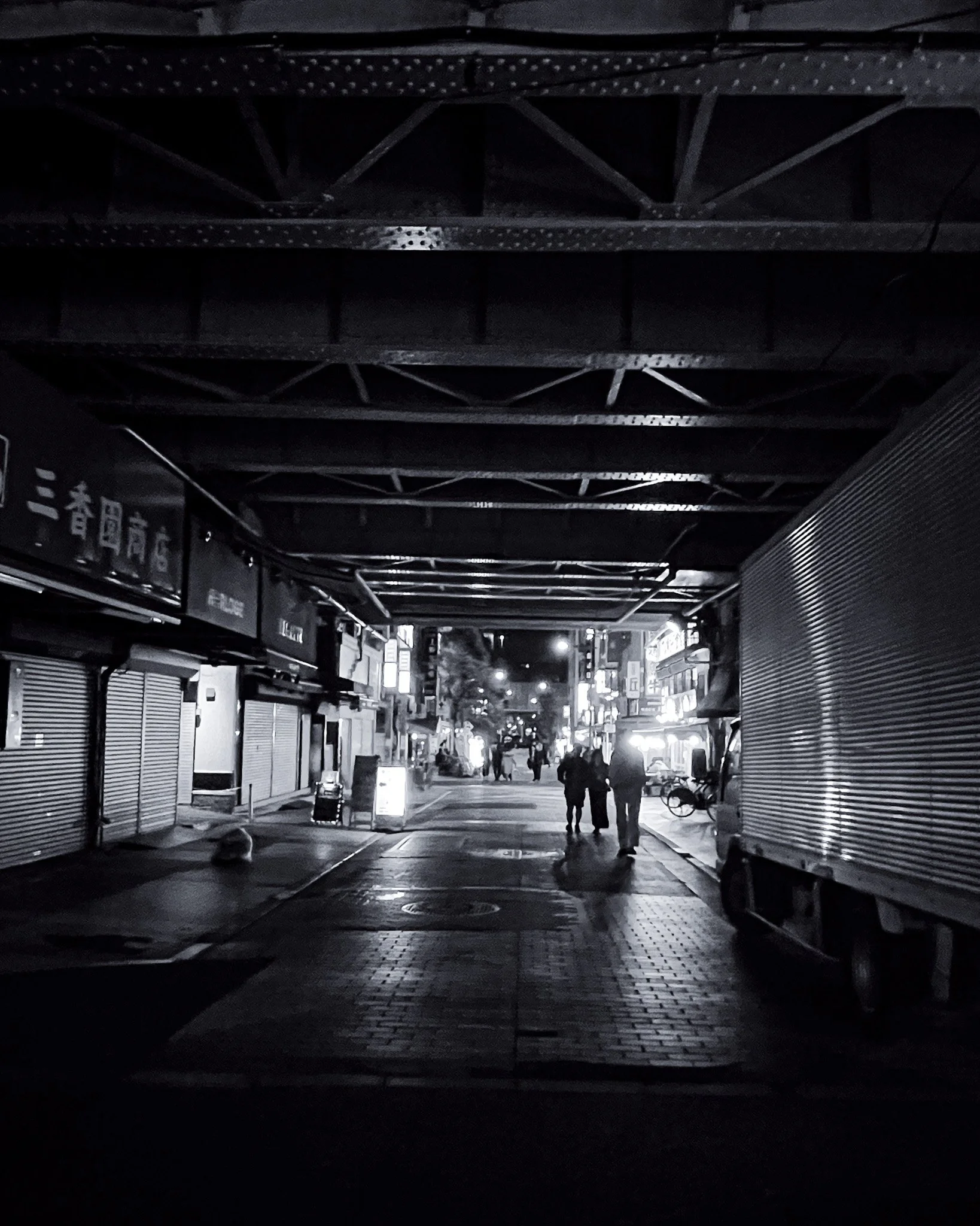 Nighttime city street viewed from underneath an overpass, with people walking and shops illuminated in the background.