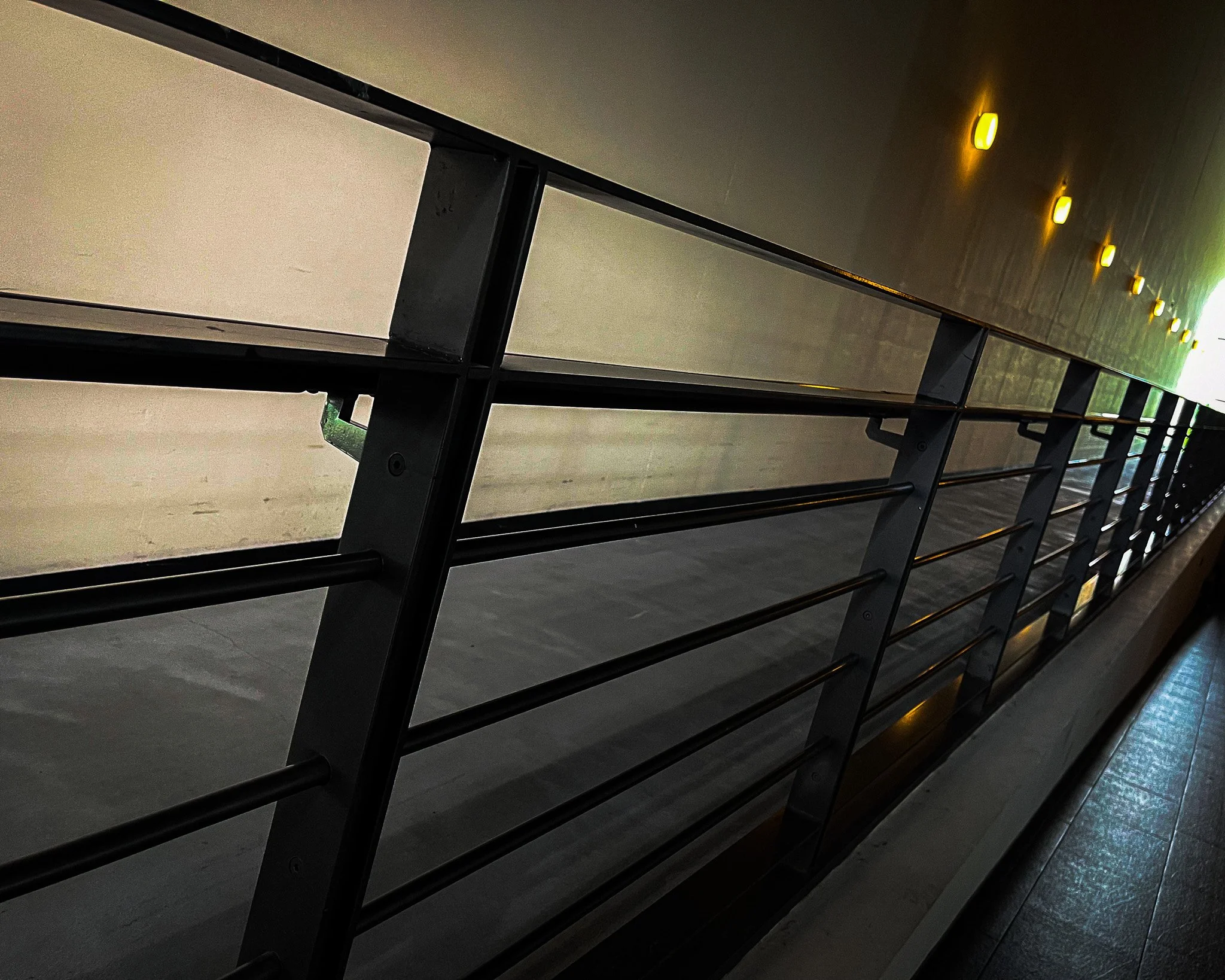 A dark corridor with a metal railing and glass panels on the left, illuminated with yellow wall-mounted lights on the right and sunlight at the far end.