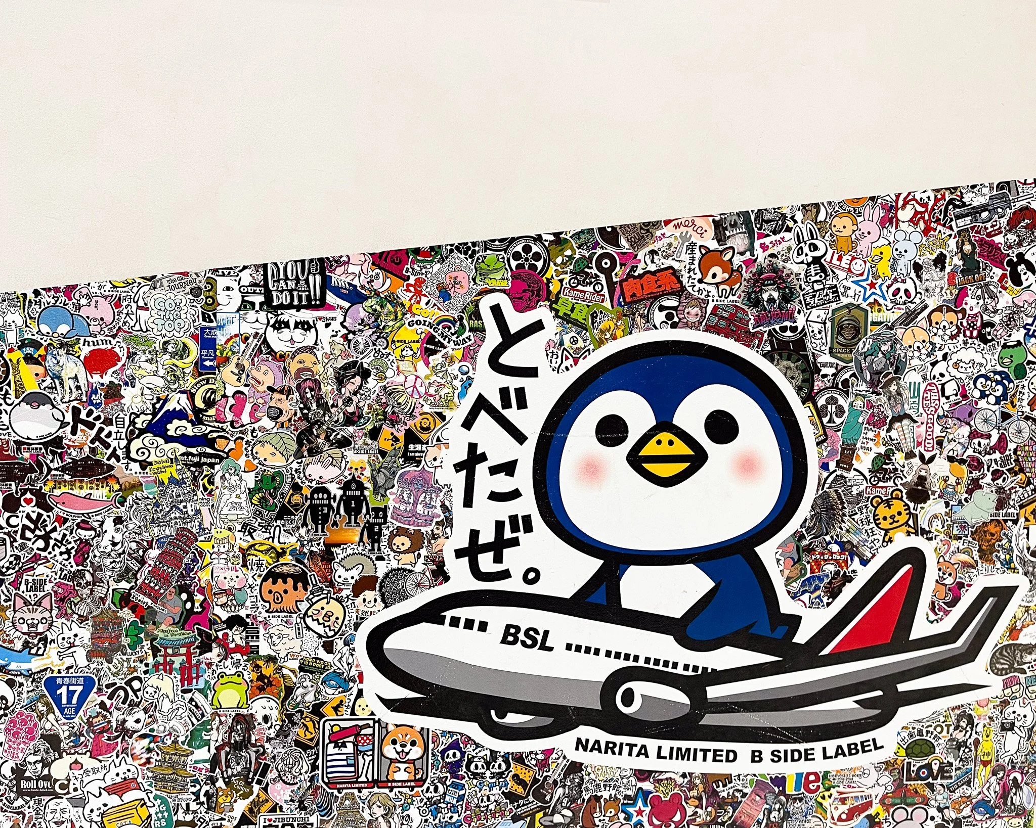 A large sticker of a cartoon penguin surfing on a surfboard with Japanese characters, with numerous smaller stickers in the background.