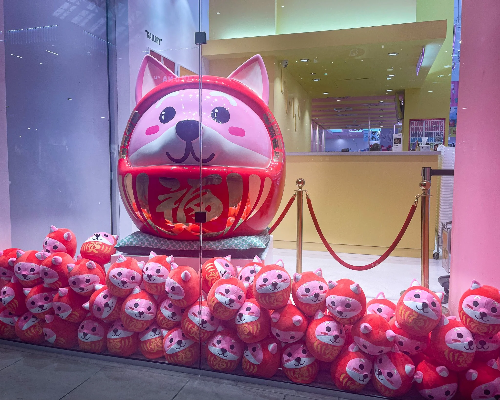 Display of many plush toys resembling the Japanese lucky cat mascot, with a large cat figure in a Daruma doll style, all in pink and red, behind glass at a shopping mall window.