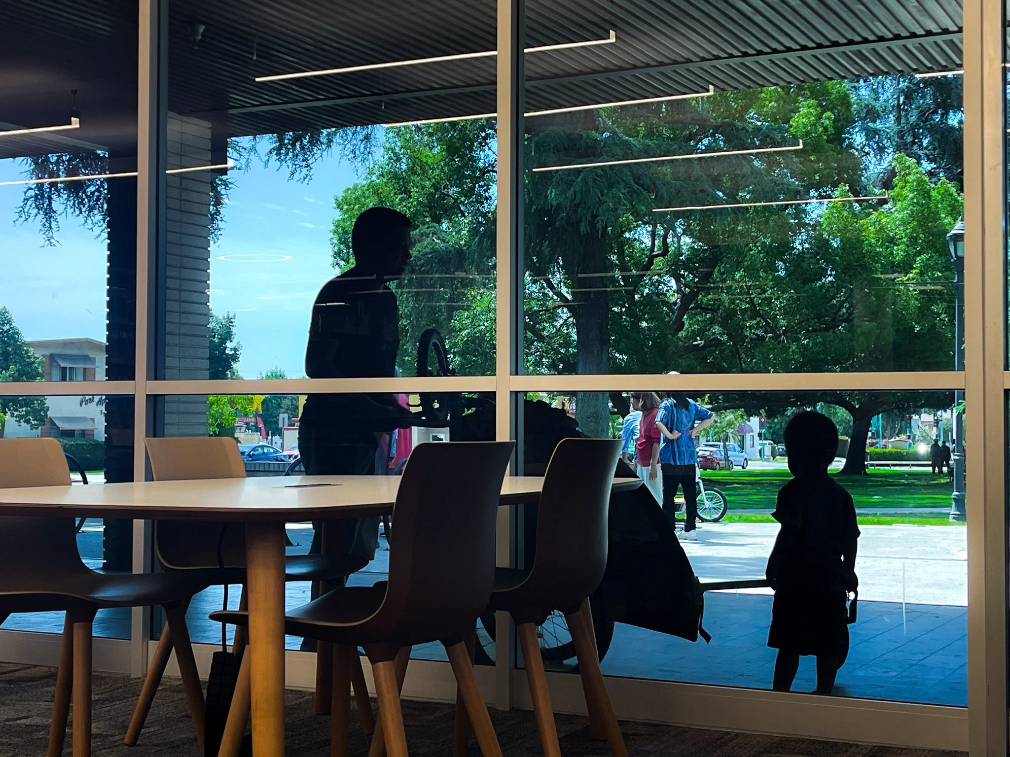 Silhouettes of a man and a child inside a building sitting near a large window, with outdoor scene of trees and people visible through the glass.