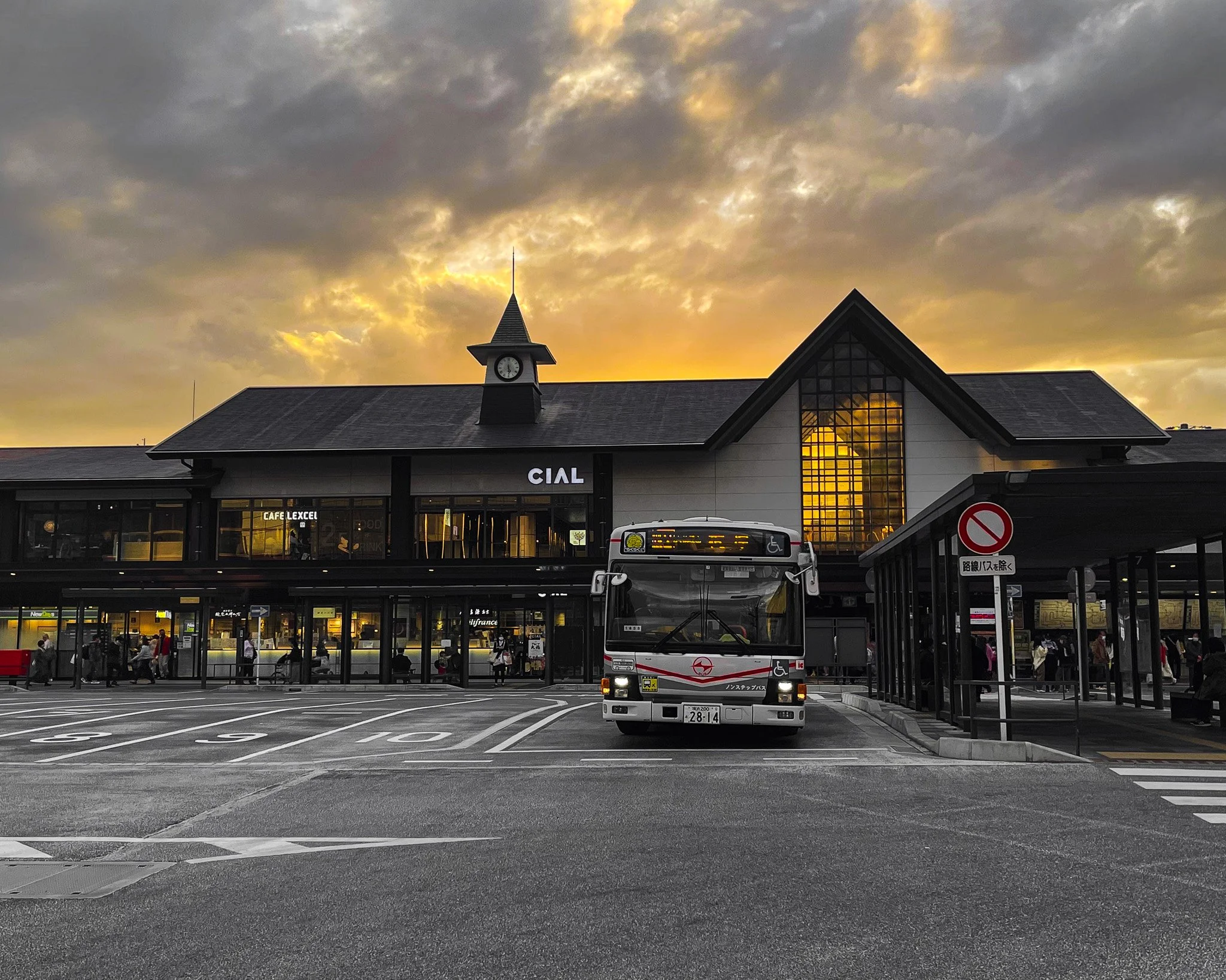 A bus parked in front of a train station during sunset, with people waiting inside and outside the station.