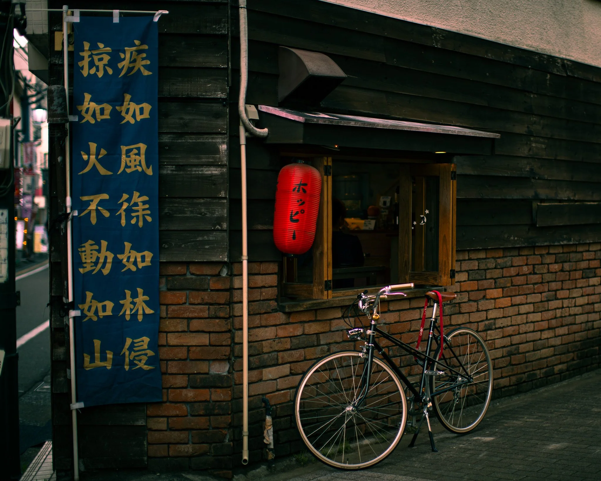 A small Japanese restaurant with a brick and dark wooden exterior, featuring a red lantern with Japanese characters, a blue vertical banner with gold Japanese characters, a window with a wooden ledge, and a black bicycle parked outside.