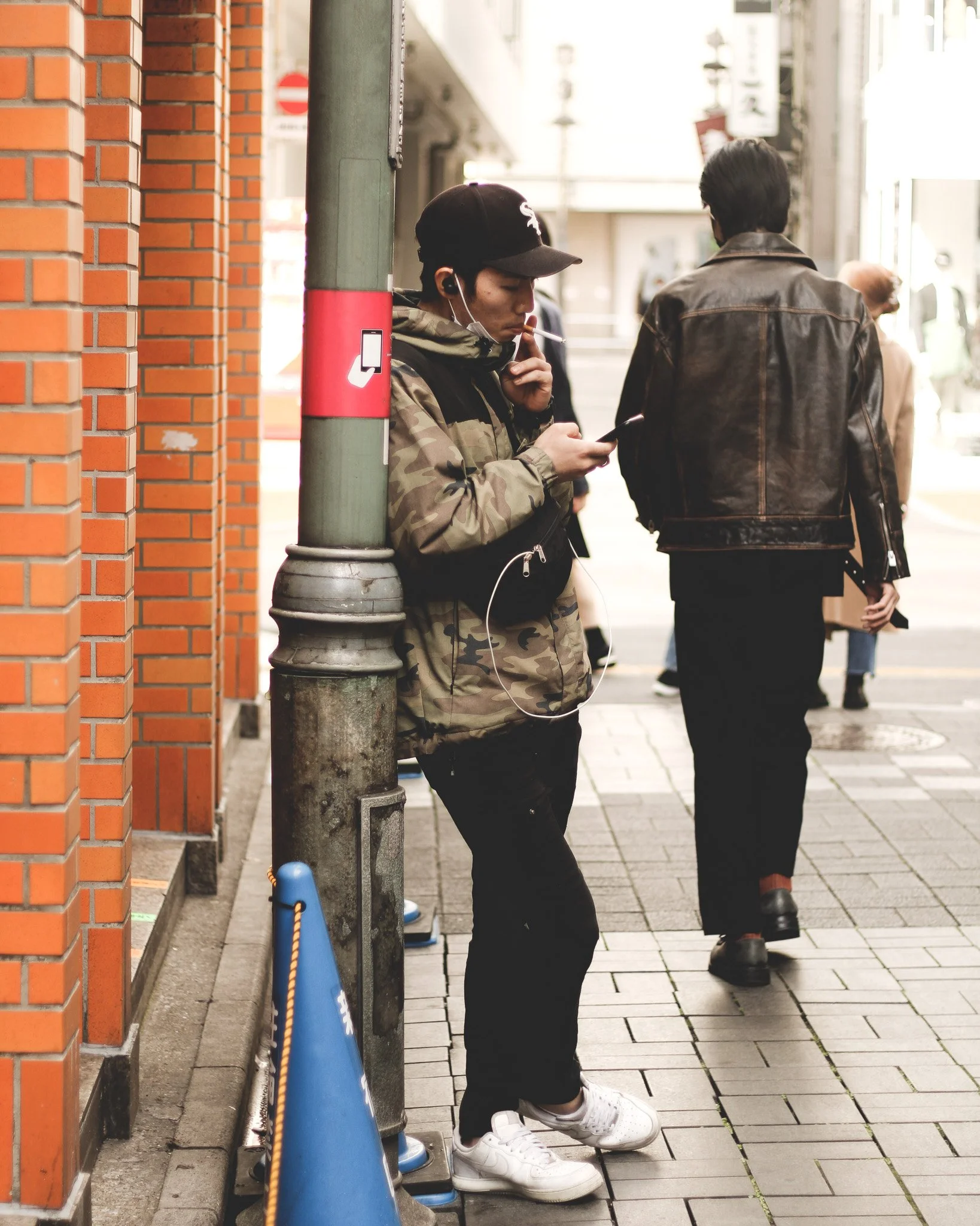 A young man with a cap, camo jacket, and earbuds is standing on a city sidewalk, leaning against a pole while looking at his phone and smoking a cigarette. He is wearing white sneakers and black pants. In the background, there are pedestrians walking