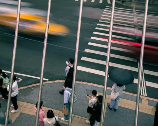 從高處欄杆間俯瞰，行人在斑馬線旁等候，黃色計程車呼嘯而過成殘影，台灣