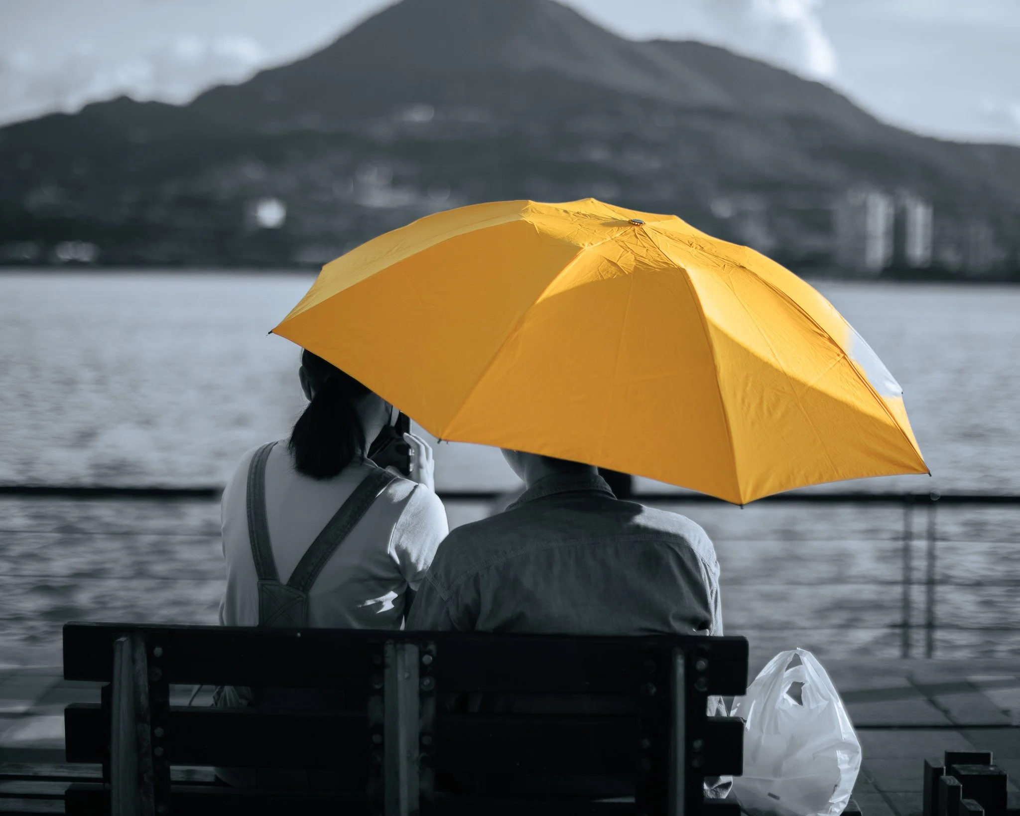 A man and woman sitting on a bench by a body of water during the day, sharing an orange umbrella. The background features a mountain and some buildings, with a cloudy sky.