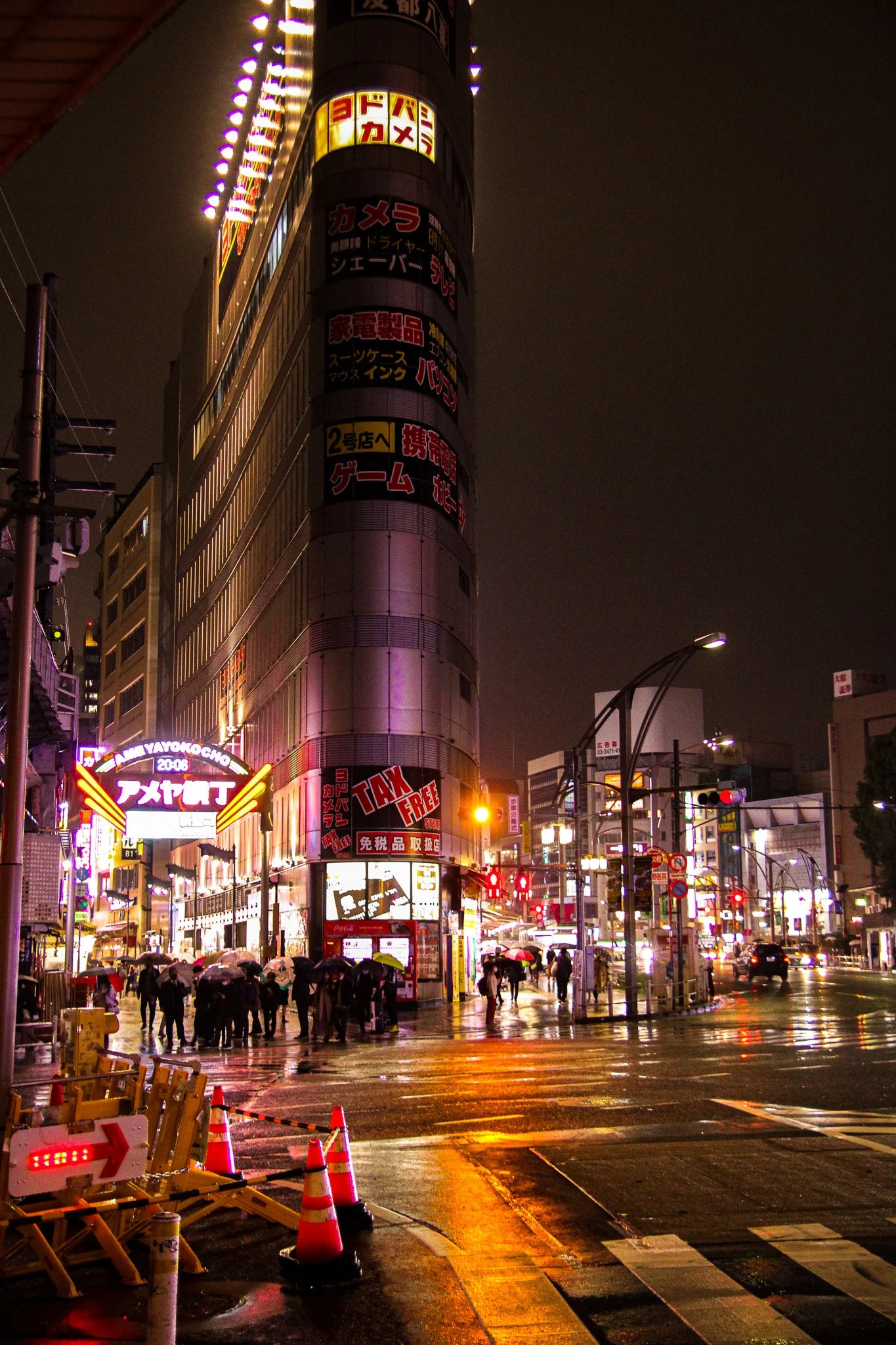 Night scene of a busy city street with illuminated signs, a tall building covered in advertisements, people holding umbrellas waiting at a crosswalk, and reflections on wet pavement.