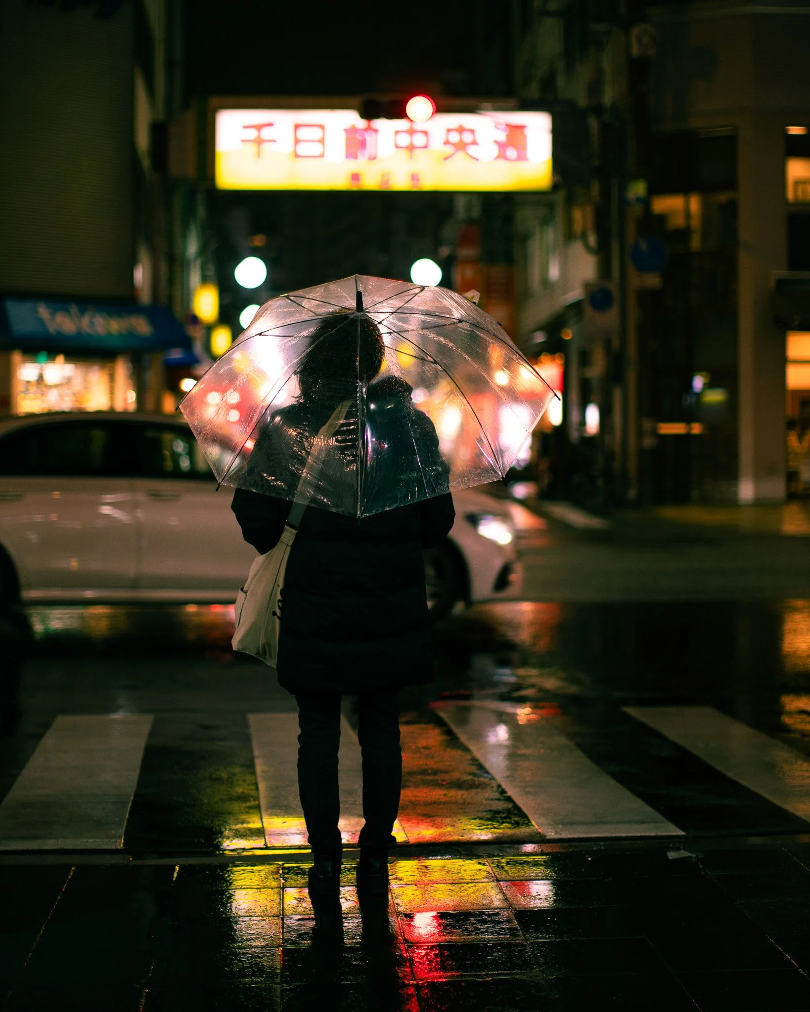Figure with a clear umbrella waiting at a rain-slicked crossing beneath neon signs at night, Osaka, 2025