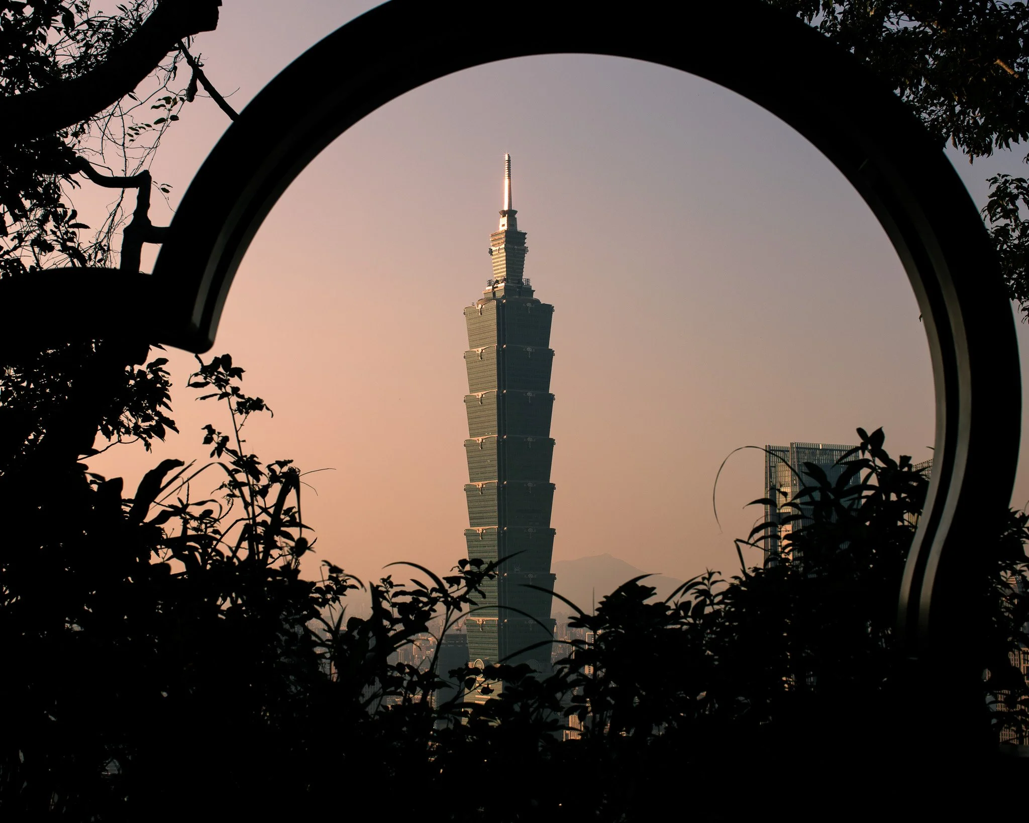 Taipei 101 skyscraper viewed through a circular frame, with silhouetted trees and plants in the foreground and a pastel sky in the background.