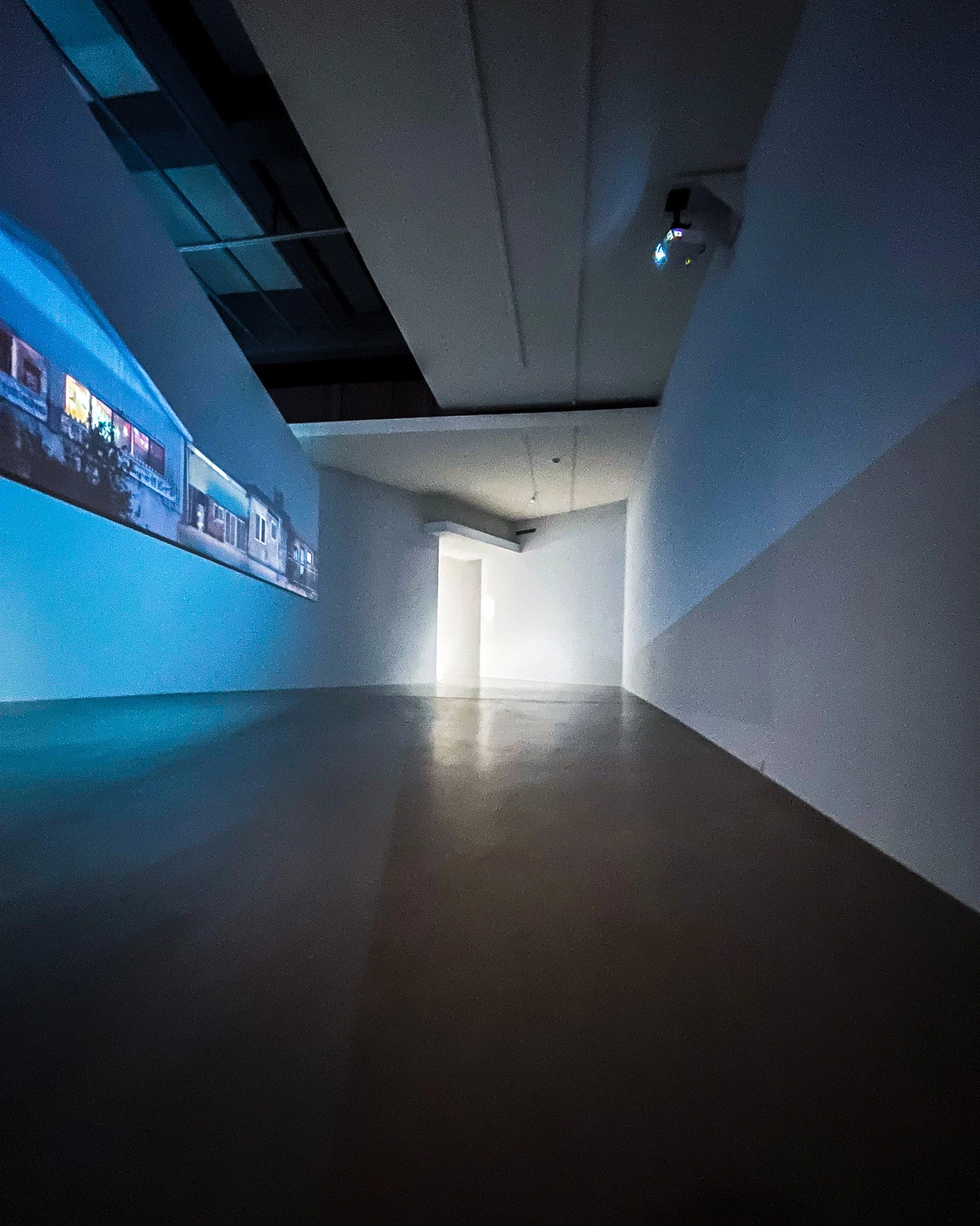 An art gallery room with a long white wall on the left displaying a projection of colorful images, lighting dim. The room has a minimalist design with a concrete floor and a small white door or opening at the back illuminated by natural light.