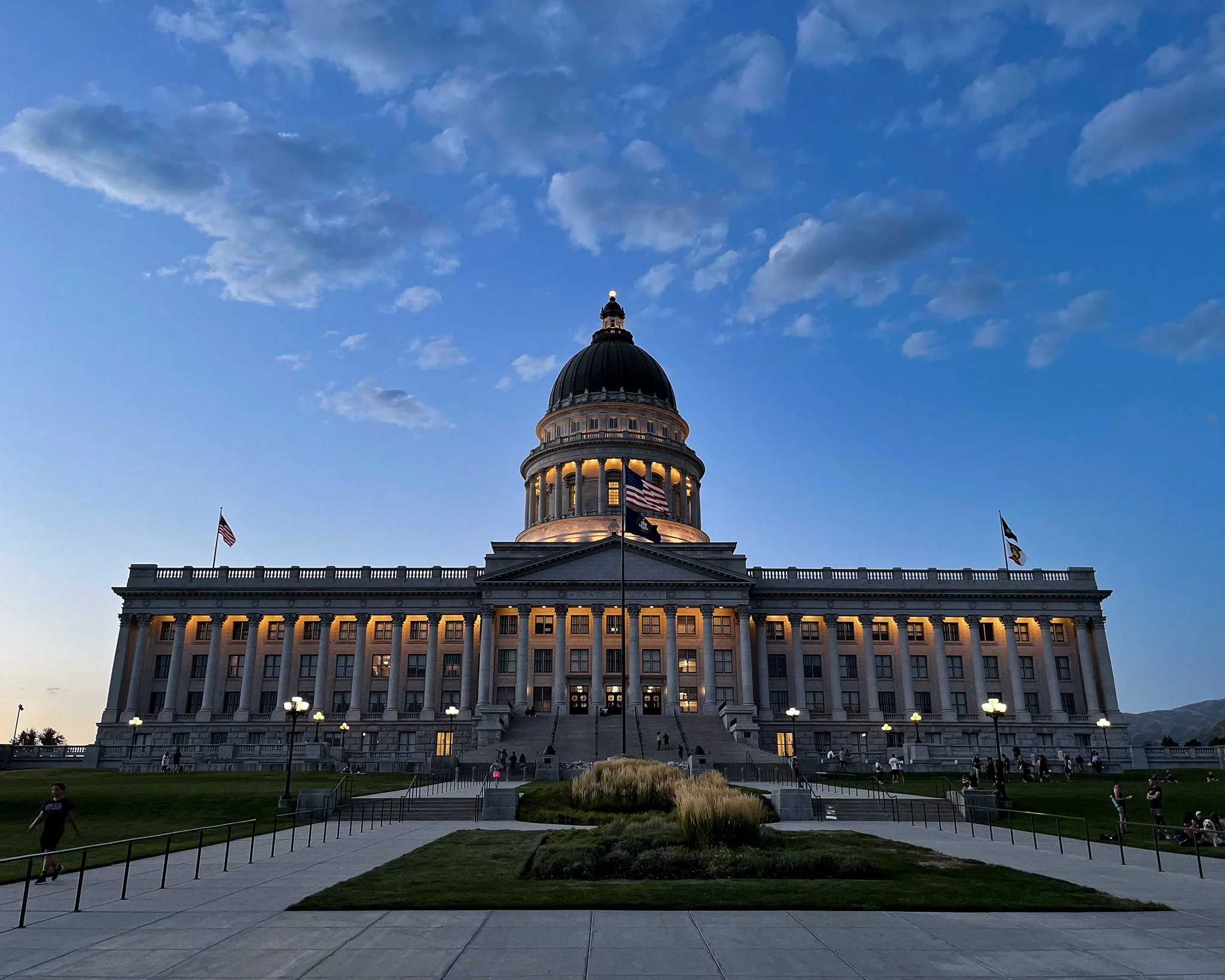 The Utah State Capitol building lit up at dusk, with a large dome, flags, and a wide staircase leading up to the entrance, surrounded by people and a landscaped area.
