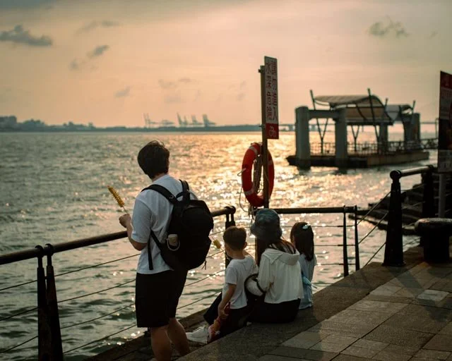 一家人聚在港邊欄杆旁的救生圈下，望著夕陽灑落水面，台灣