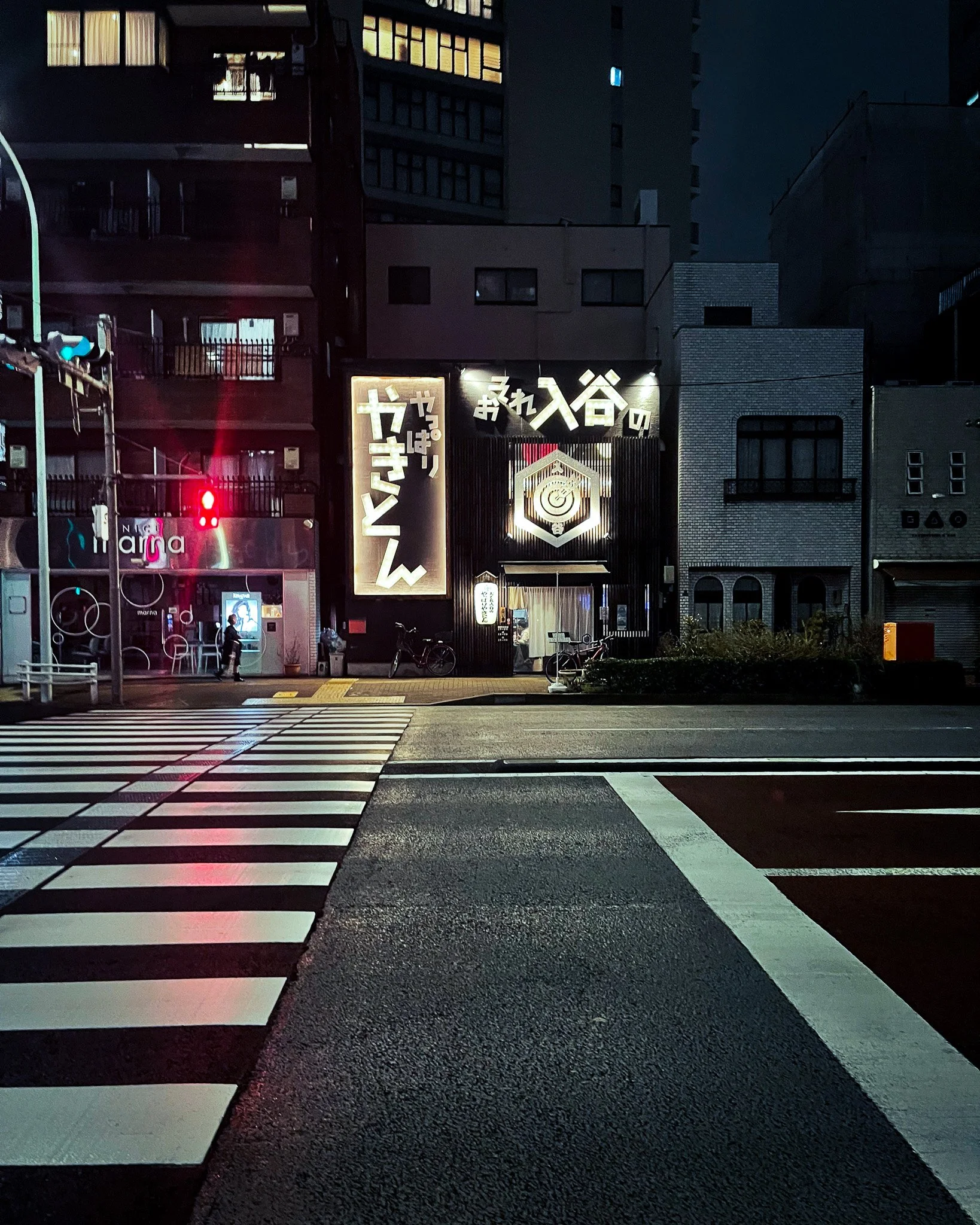 Nighttime city scene with a crosswalk in the foreground, a traffic light showing red, and buildings with illuminated signs in the background, including Japanese characters.