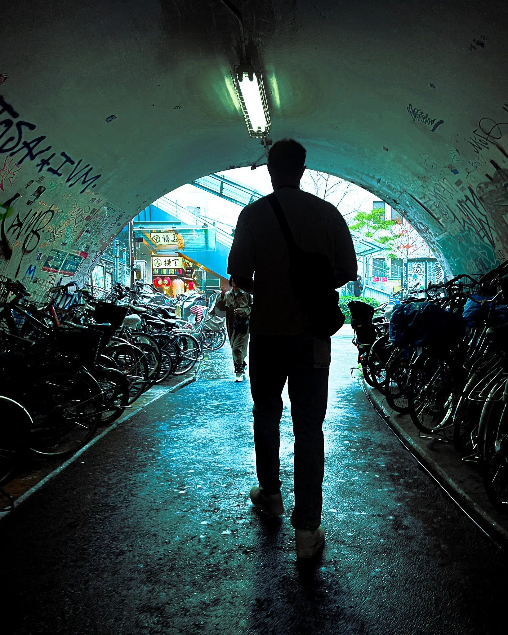 Silhouette of a person walking through a graffitied underpass, with bicycles parked on both sides and an outdoor area visible at the end of the tunnel.