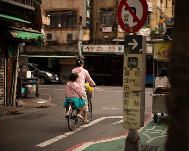 兩人共乘一輛腳踏車，經過松山車站指示牌旁的路口，台北