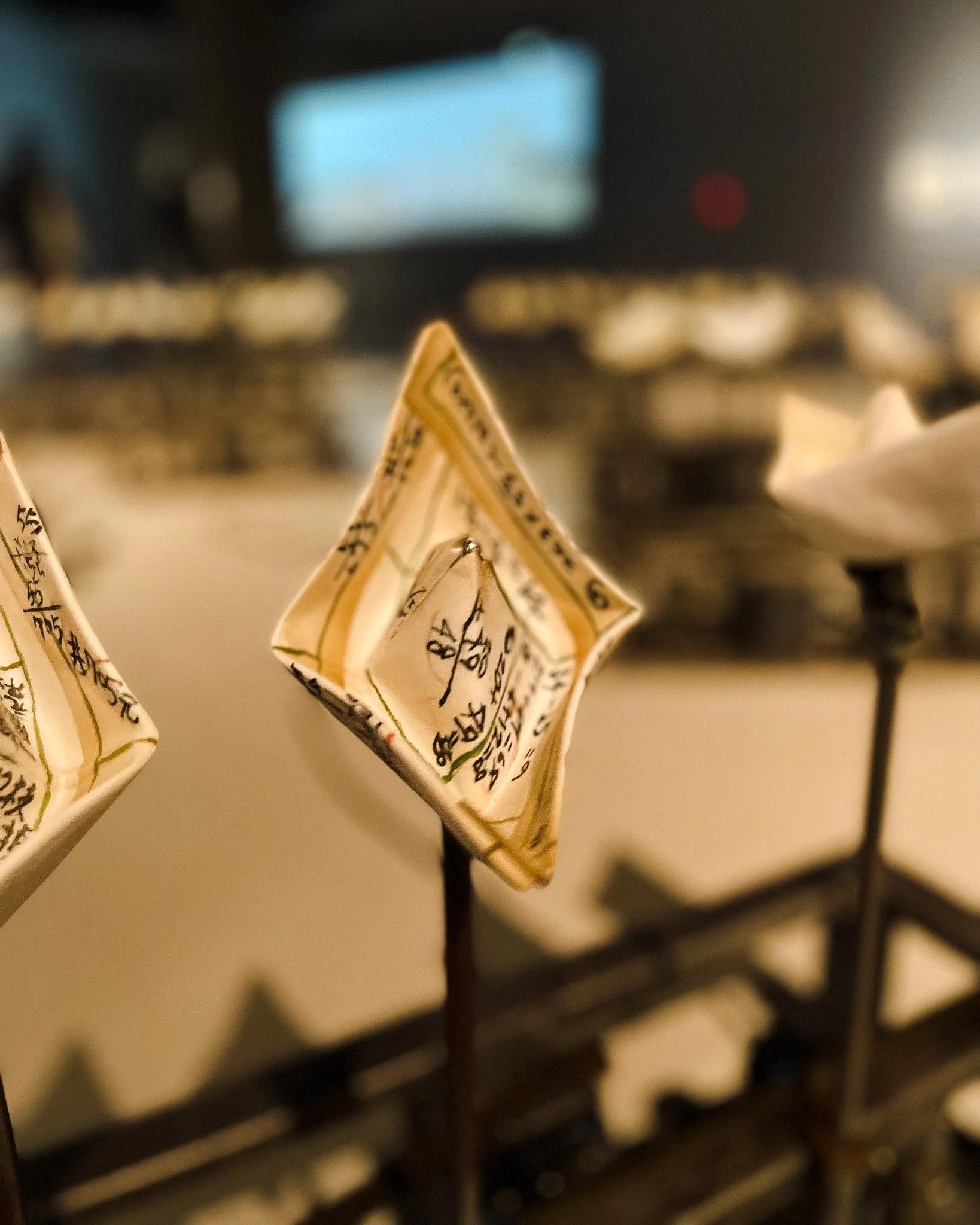 Close-up of a folded paper coin with handwritten notes, displayed on a stand, with blurred background of a cash register and a blurred screen.