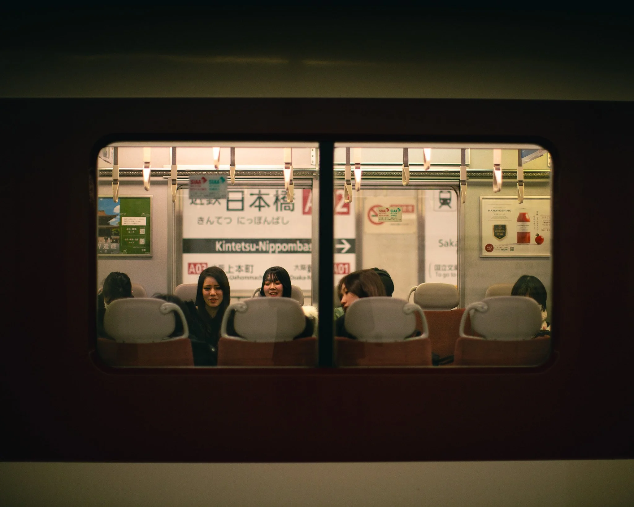 Passengers seated inside a train carriage seen through the window, with Japanese signage above