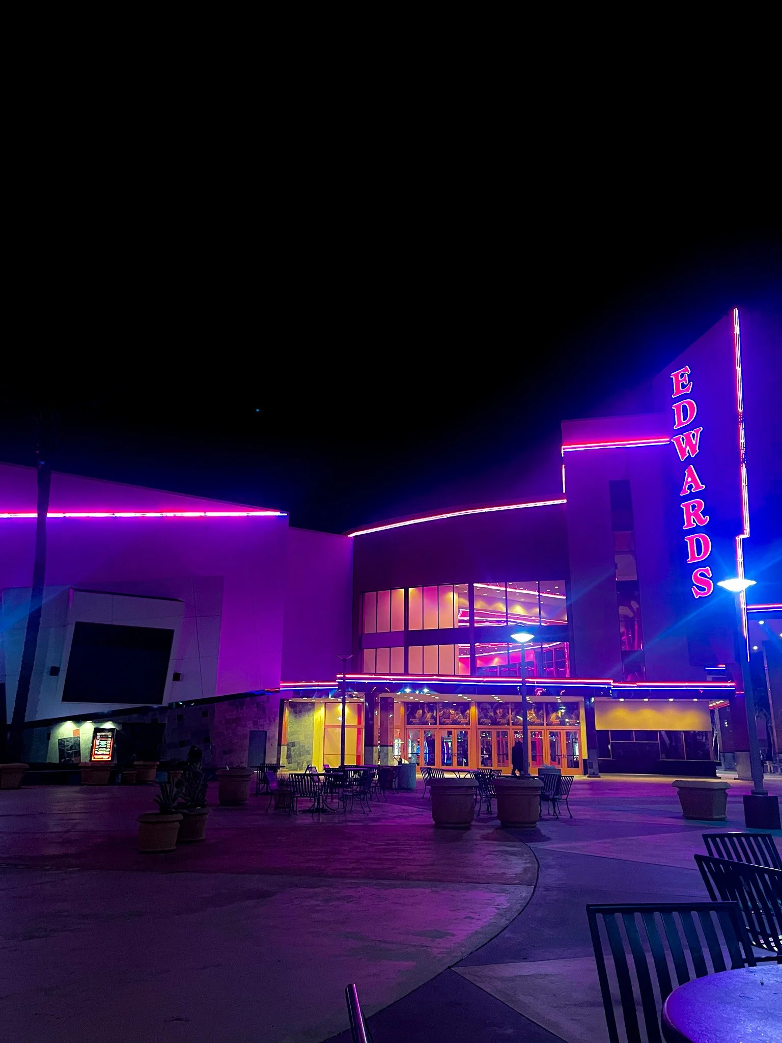 Night view of a brightly lit entertainment venue called EDWARDS with neon purple and pink lights, outdoor seating, and a modern architectural design.