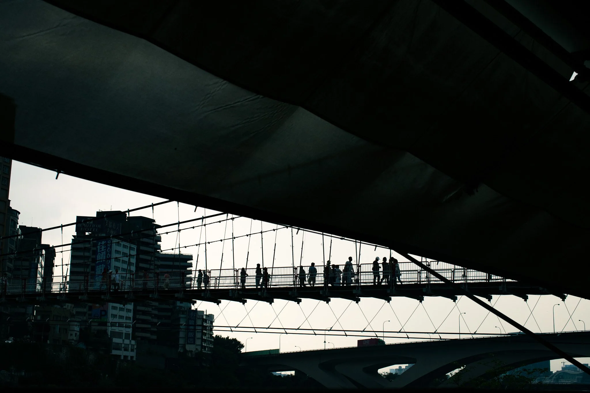 Silhouettes of people walking on a suspended bridge against a cloudy sky in an urban setting.