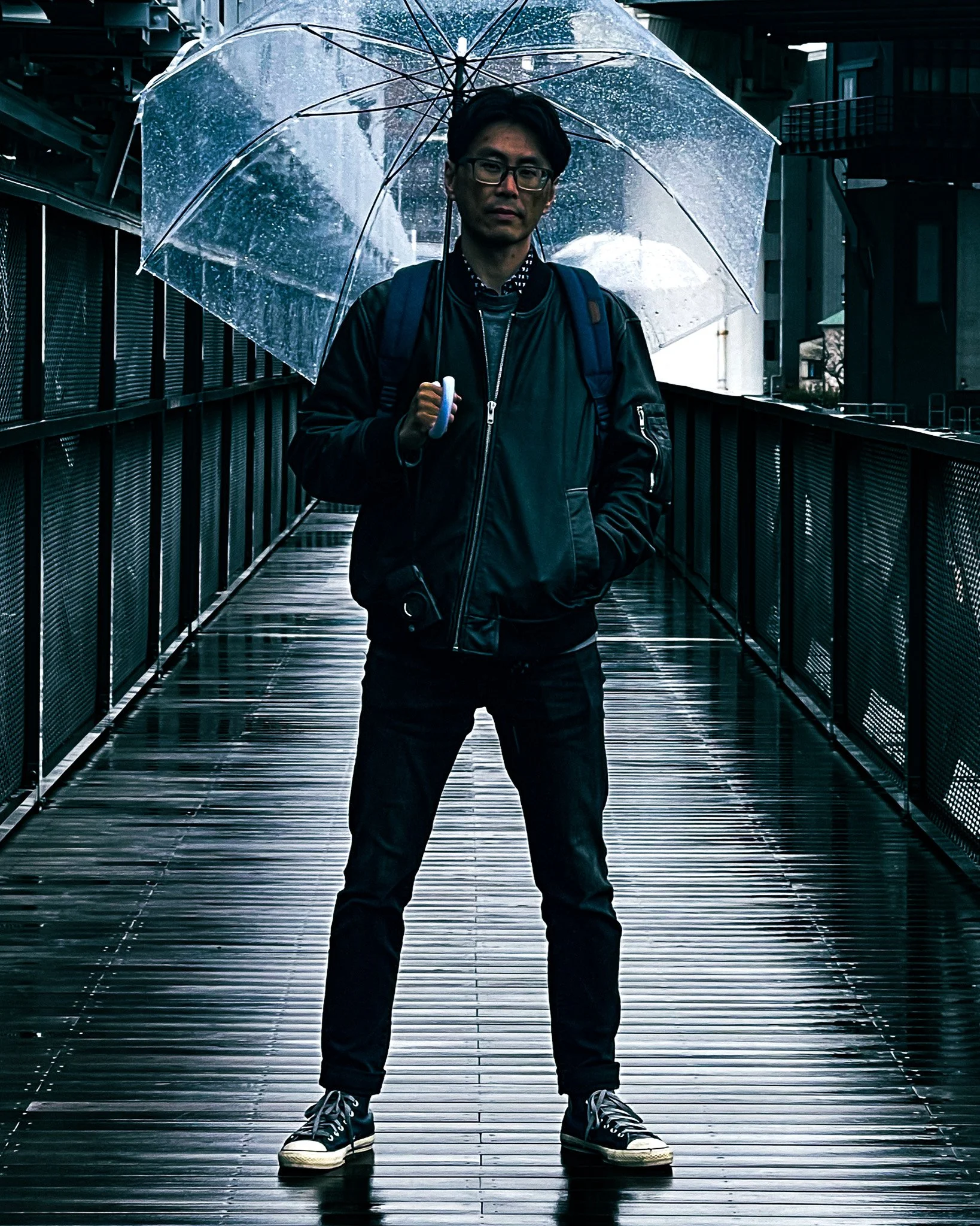 A man standing on a wet wooden bridge holding a transparent umbrella in rainy weather.