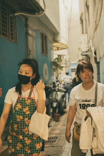 兩名女子走過藍牆與機車夾道的窄巷，其中一人繫著花圍裙，台灣