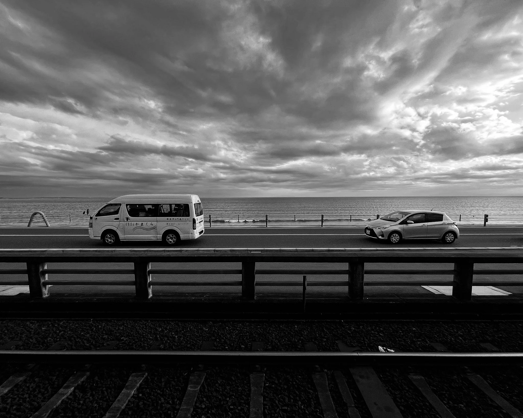 Black and white photo of a road with a van and a small car parked, with an ocean and cloudy sky in the background, and train tracks in the foreground.