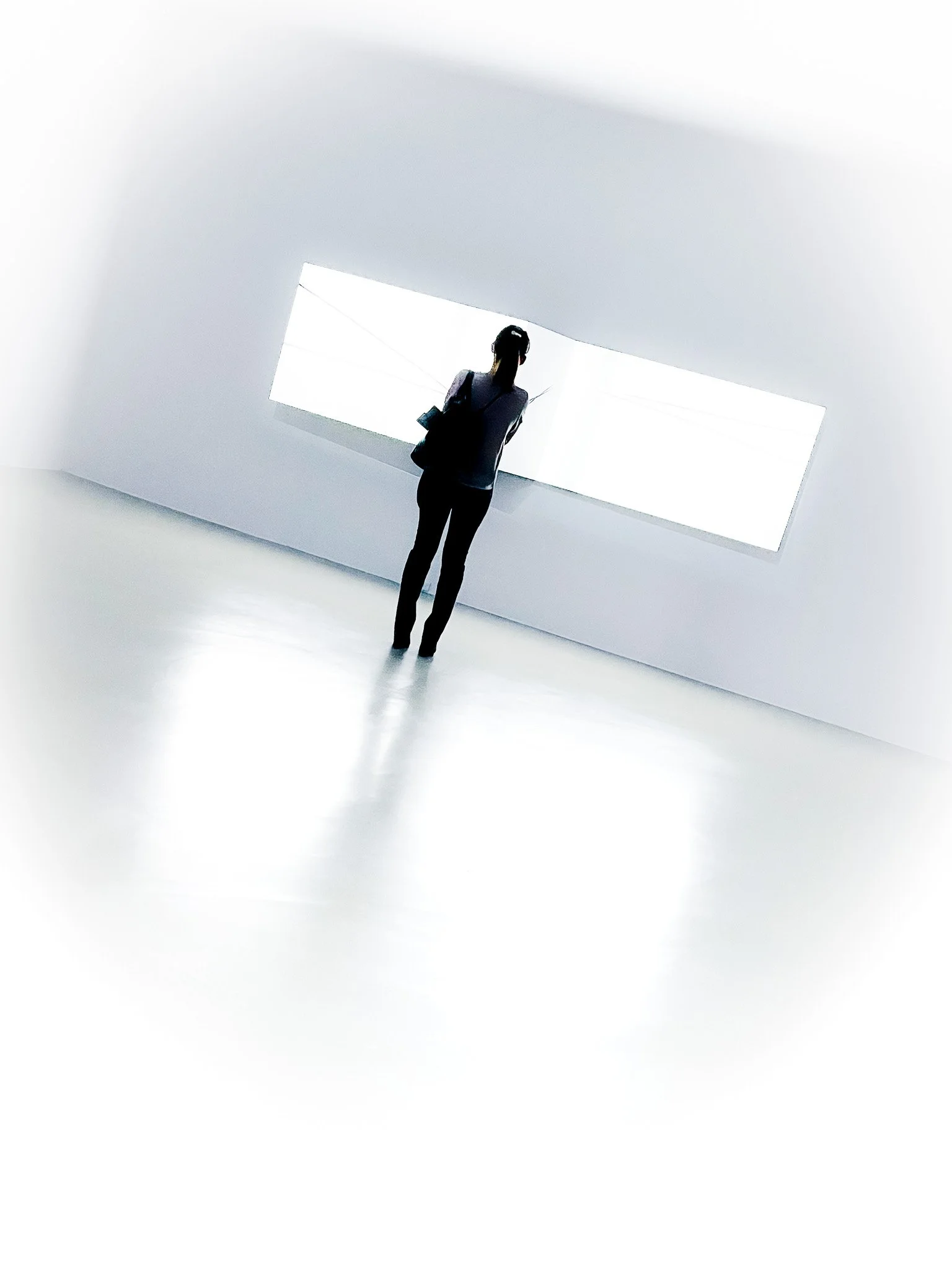 A person with a ponytail, dressed in dark clothes, stands in front of a white-lit rectangular display in a minimalist, white room, viewing the display.