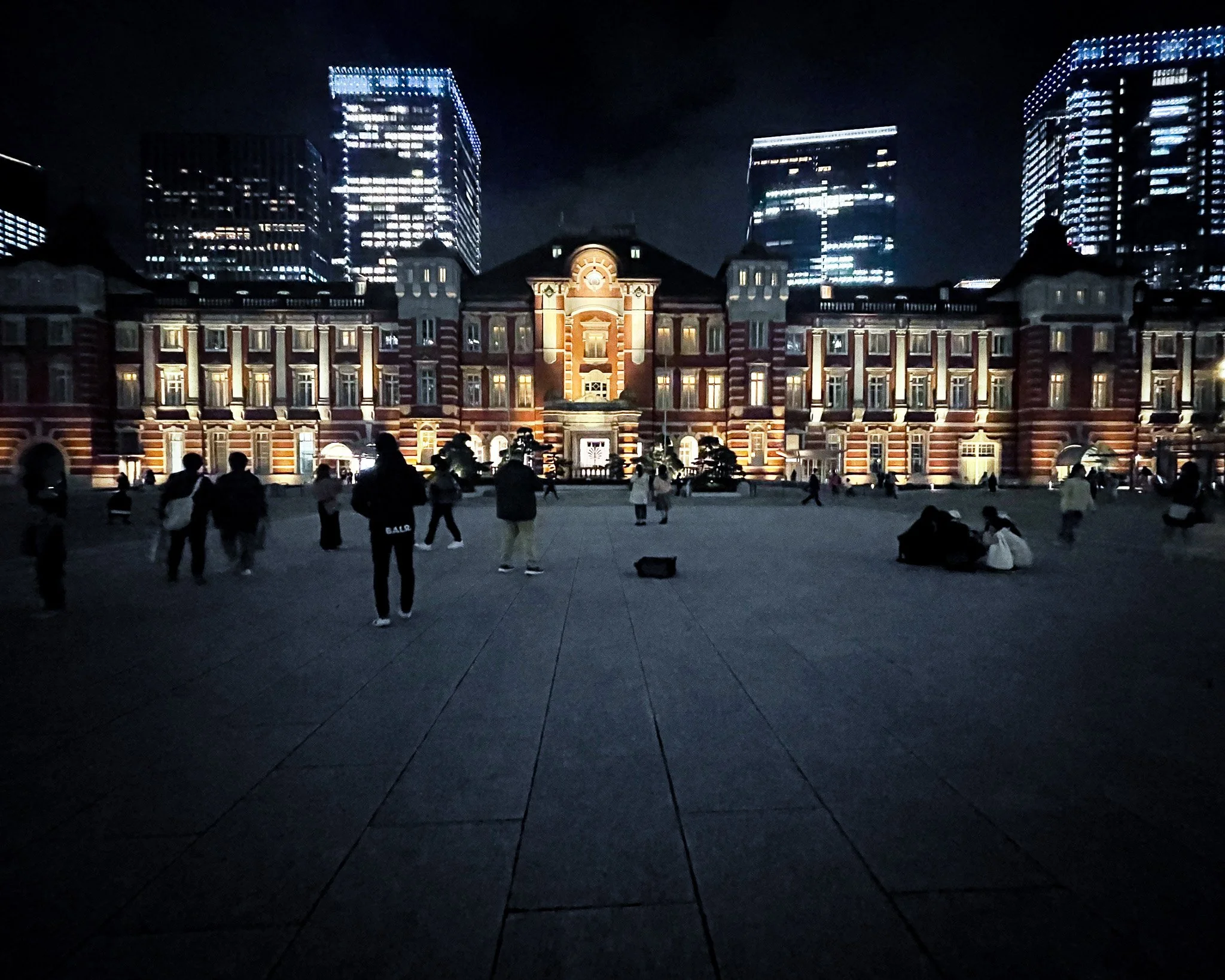 Night view of a historic red brick building with illuminated windows and modern high-rise buildings in the background, people walking and sitting in a large open plaza.